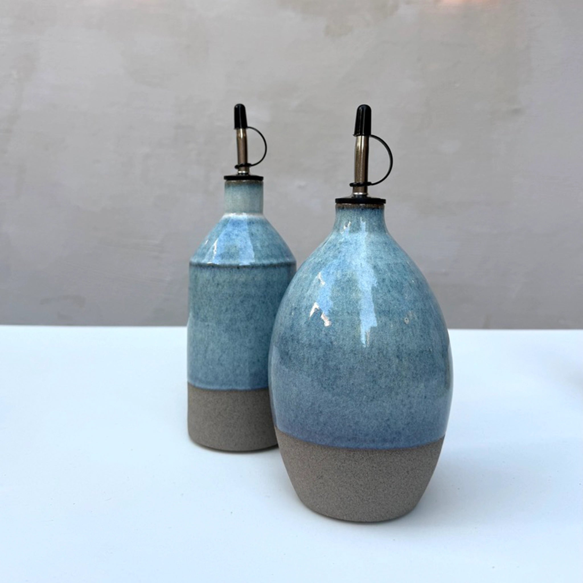 Twee AB Ceramics Olijfolie Fles Grijs Opaal 84 cl dispensers, elk handgegoten met een blauw en grijs glazuur en metalen schenktuiten, zijn afgebeeld op een wit oppervlak tegen een lichtgrijze achtergrond.