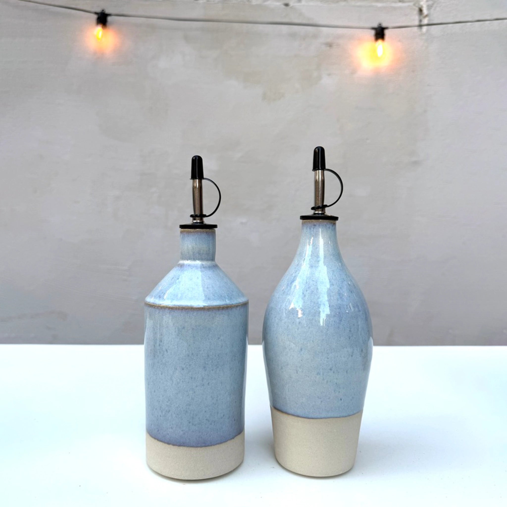 Twee AB Ceramics Olijfolie Fles Wit Opaal 54 cl flessen met metalen tuiten en beige bodems staan naast elkaar op een wit oppervlak, verlicht door twee zachte, warme strijklichtjes op de achtergrond.