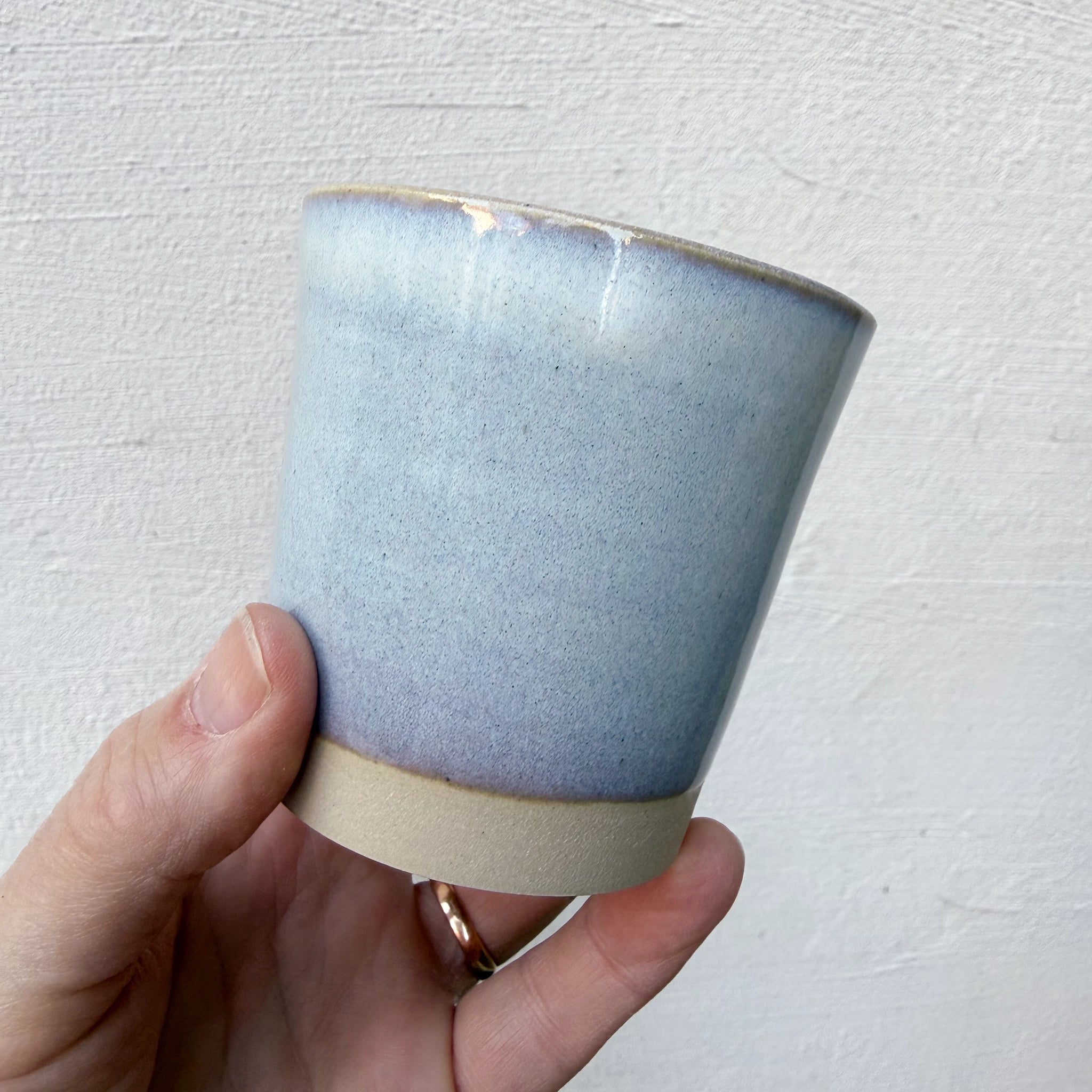 Een hand houdt de AB Ceramics Beker Wit Opaal 20 cl vast, met een mat lichtblauw glazuur bovenop en een ongeglazuurde lichtbeige witte steengoed klei basis, tegen een witte achtergrond met textuur.
