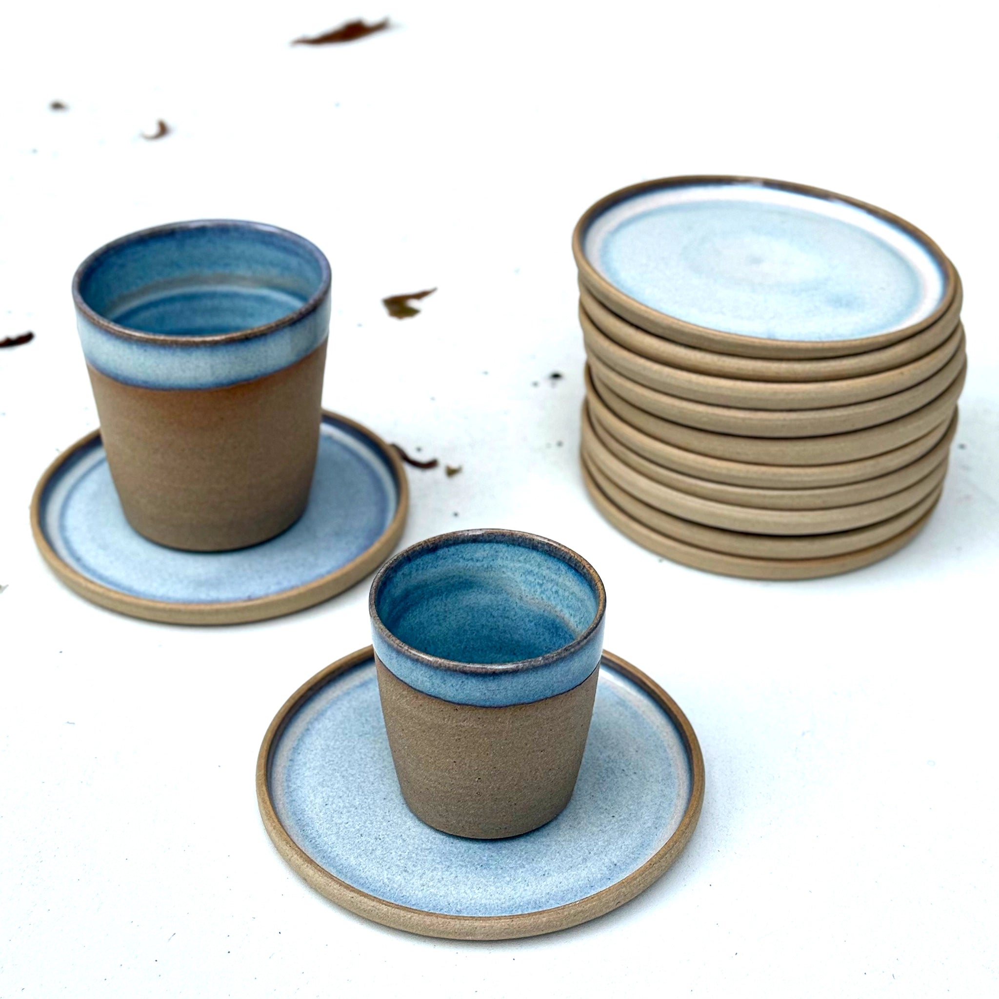 Twee AB Ceramics Espressokopjes Geel Opaal/Blauw 10 cl zitten naast een stapel borden. De handgemaakte kopjes hebben lichtblauw glazuur en bruine randen, gerangschikt op een wit oppervlak met verspreide bladeren.