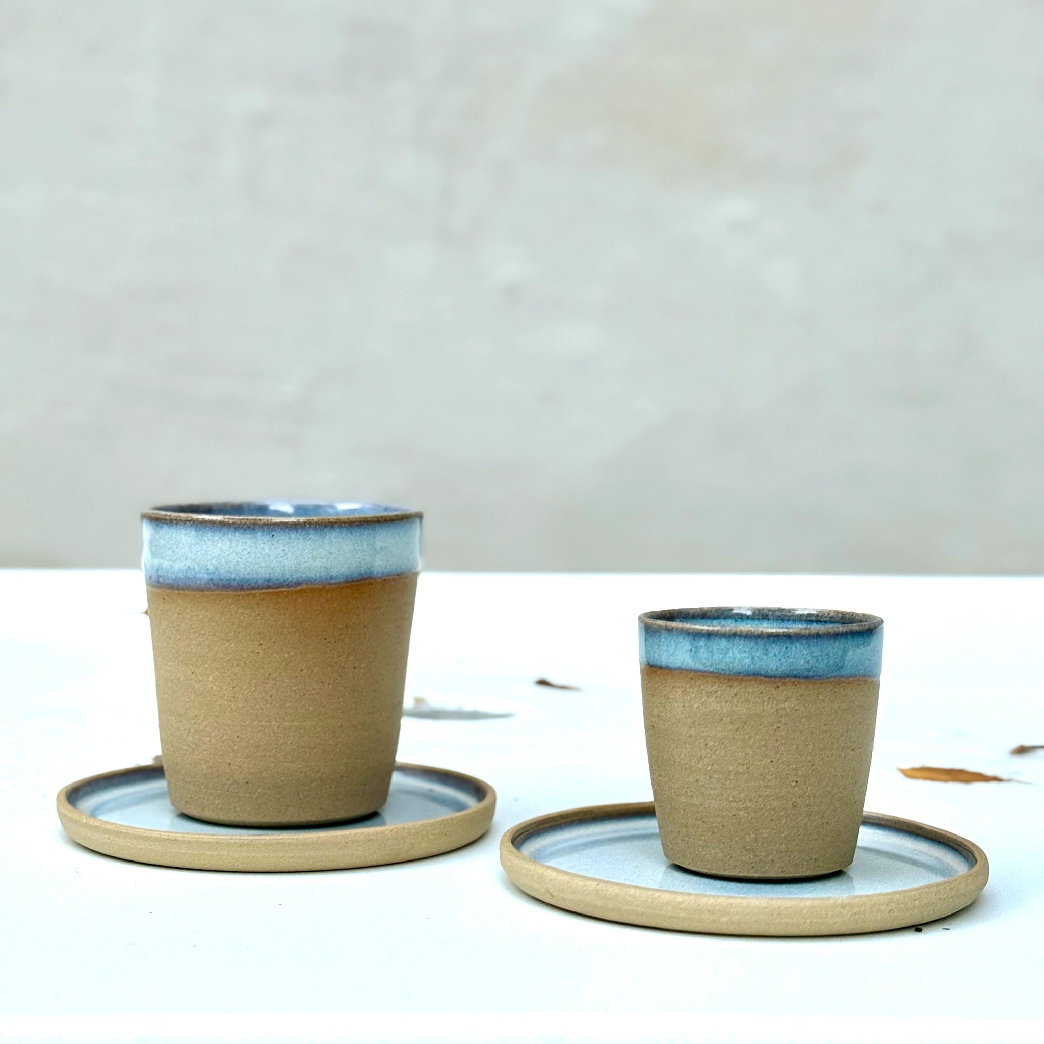 Twee AB Ceramics Espressokopjes Geel Opaal/Blauw 10 cl, gemaakt van gele steengoedklei met glanzend blauwe randen, staan samen met schoteltjes op een effen wit oppervlak. De achtergrond is eenvoudig en neutraal.