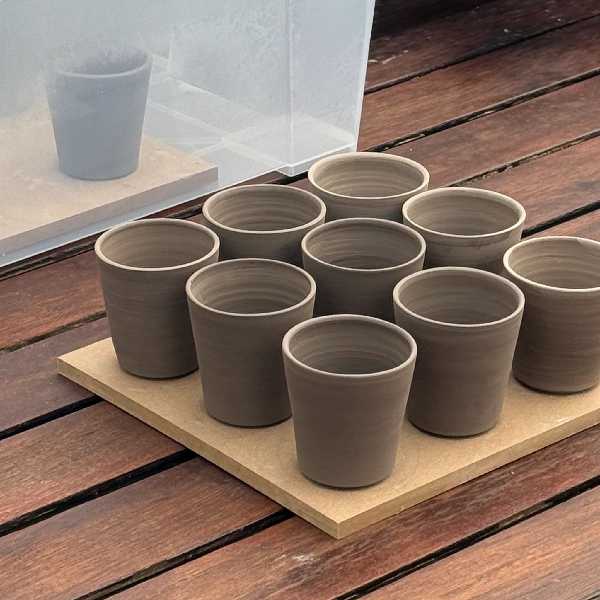 Negen ongeglazuurde kopjes van klei, handgemaakt in Antwerpen, staan op een houten plank. Daarachter wordt de AB Ceramics Beker Toffee Satijn 20 cl weerspiegeld in een doorschijnende doos.
