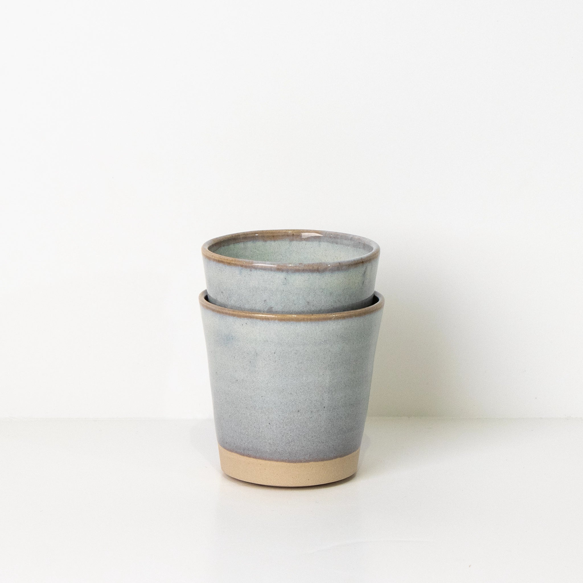 Twee AB Ceramics Beker Wit Opaal 20 cl, handgemaakt en -gedraaid van wit steengoed klei met lichtgrijze glazuur en ongeglazuurde beige voet, gestapeld op een witte ondergrond tegen een neutrale achtergrond.