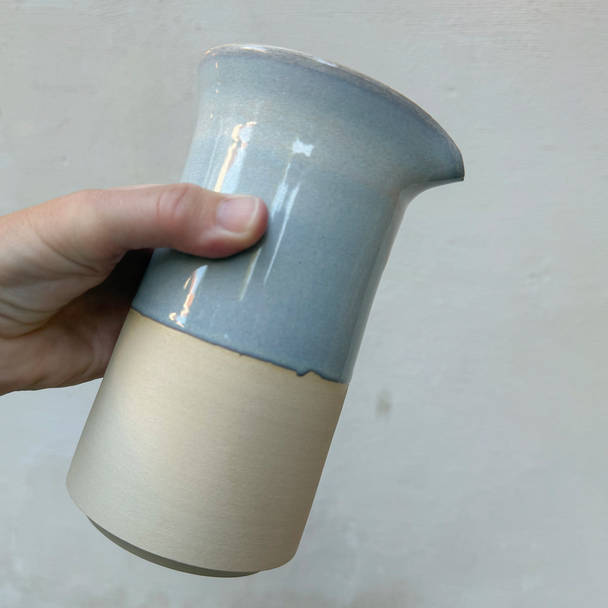 Een hand houdt de AB Ceramics Karaf Prai Opaal 65 cl schuin vast tegen een grijze muur.