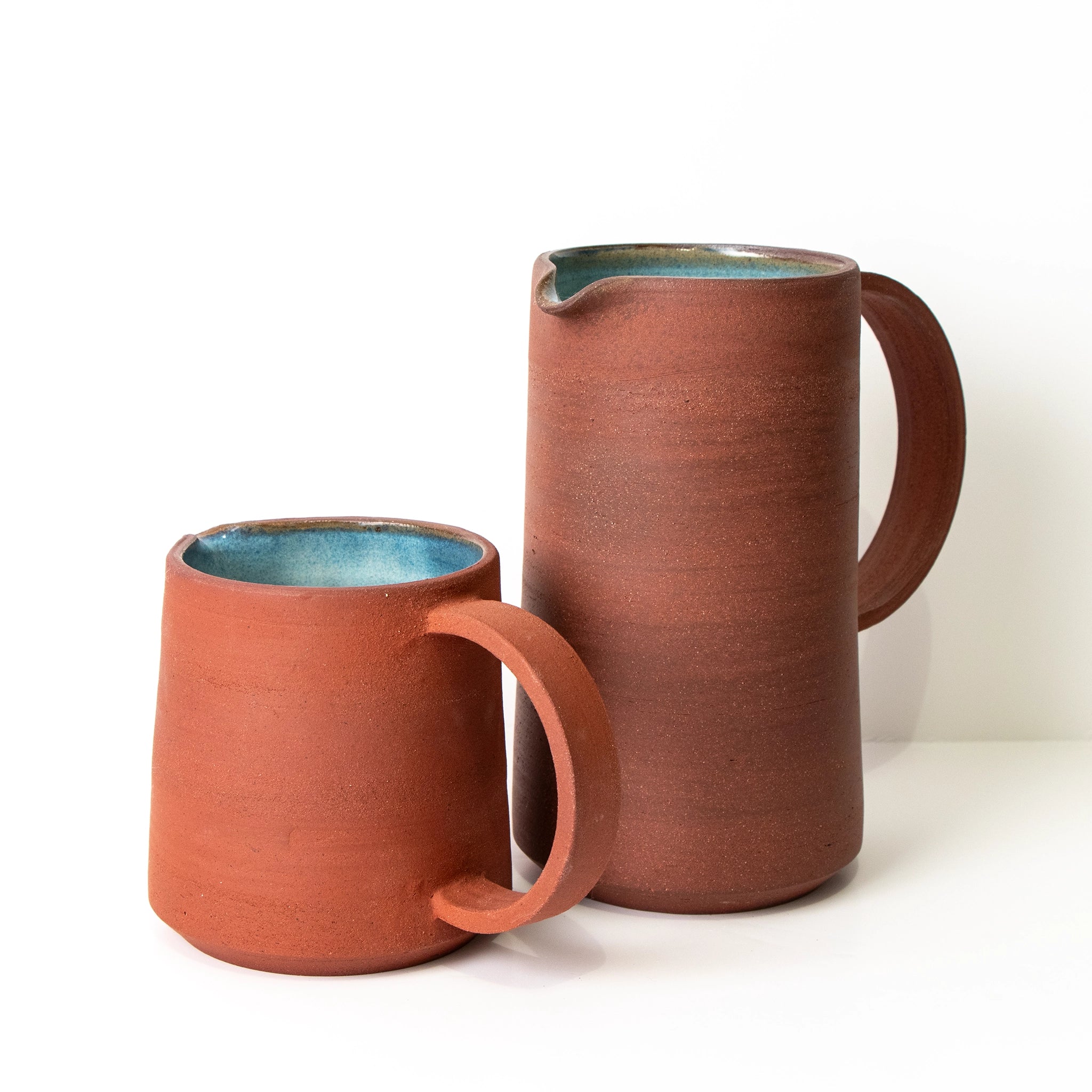 De AB Ceramics Karaf 9 Rood Opaal Blauw 40 cl en bijpassende mok zijn gemaakt van rode klei met een rustieke roodbruine buitenkant en blauwgroen opaalglazuur aan de binnenkant, beide op een witte achtergrond.