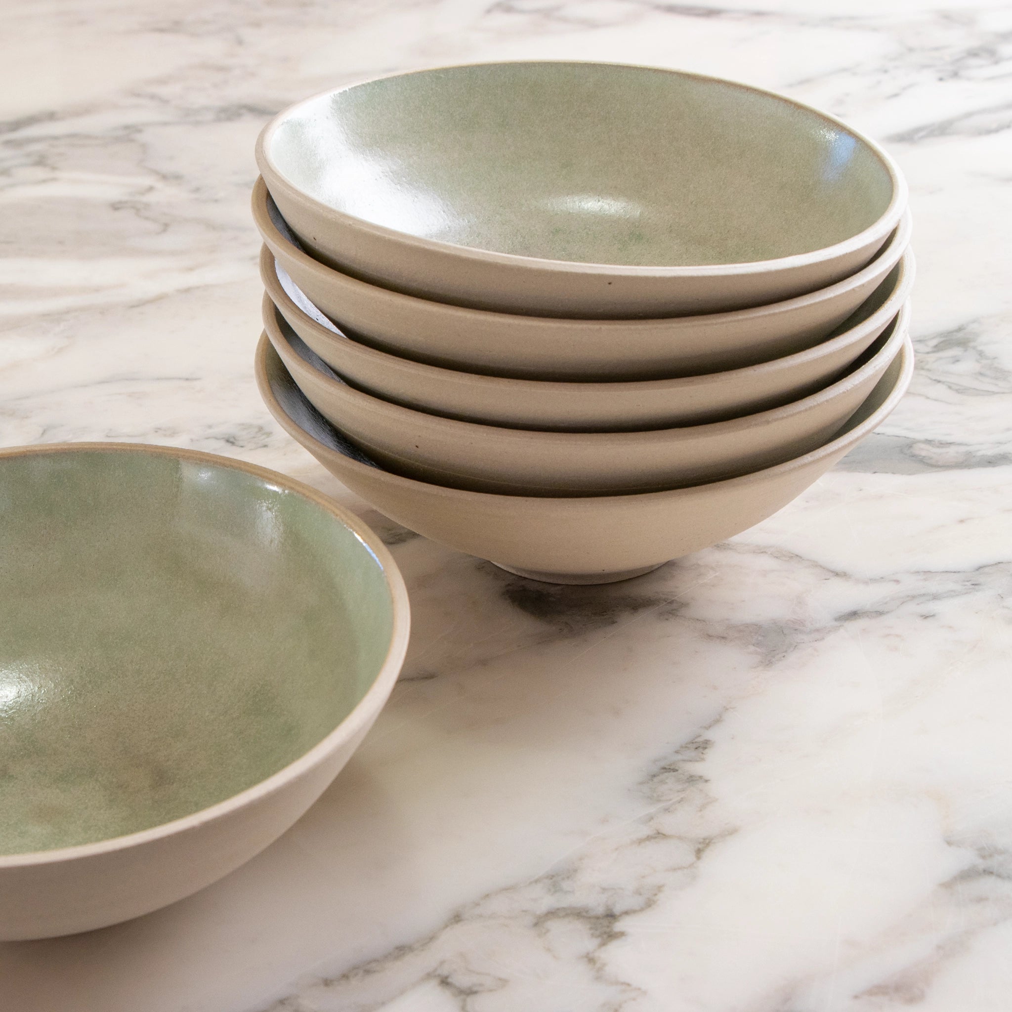 Een stapel van vijf AB Ceramics Noodle Kom Wit Mint D19 kommen met een groenige binnenkant staat op een marmeren aanrecht, met nog een AB Ceramics handgemaakte noedelkom naast de stapel.