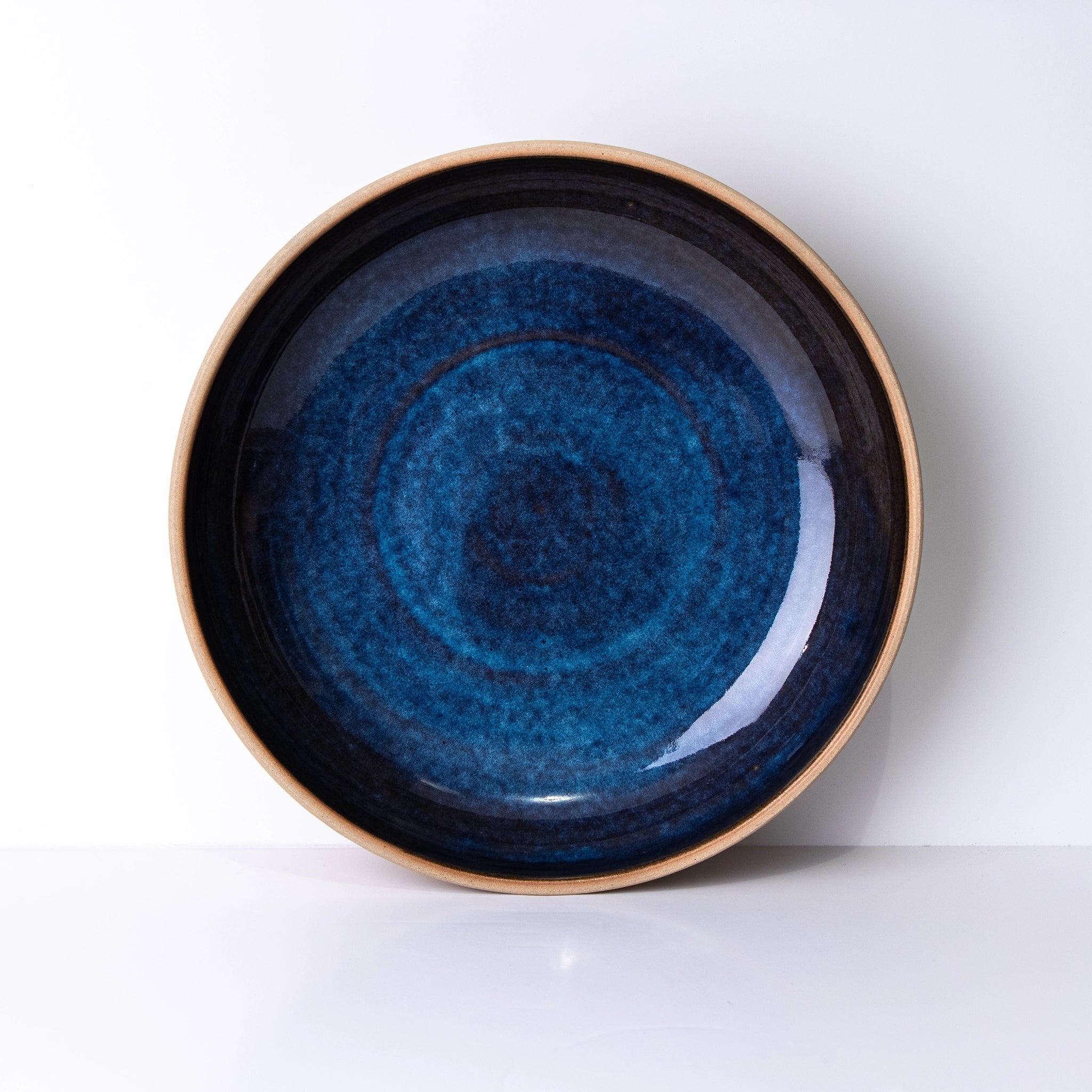 De AB Ceramics Serveerschaal Geel Blauw D25 is een ronde steengoed schaal met een glanzend diepblauw centrum en een natuurlijke bruine rand, afgebeeld tegen een effen witte achtergrond.