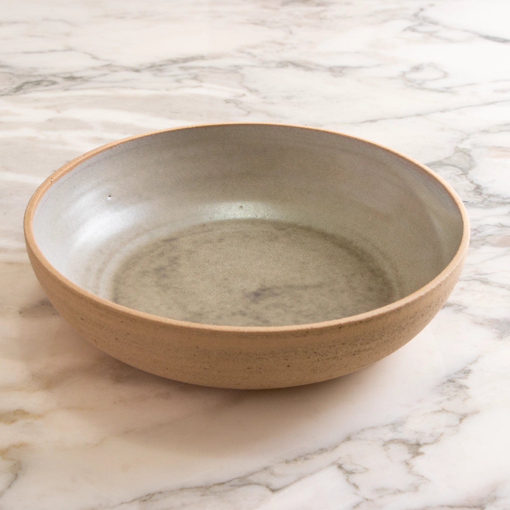 De AB Ceramics Serveerschaal Ruw Mint D27,5 is een ronde, ondiepe serveerschaal met een matbeige buitenkant en een gladde, lichtgrijze binnenkant, weergegeven op wit marmer.