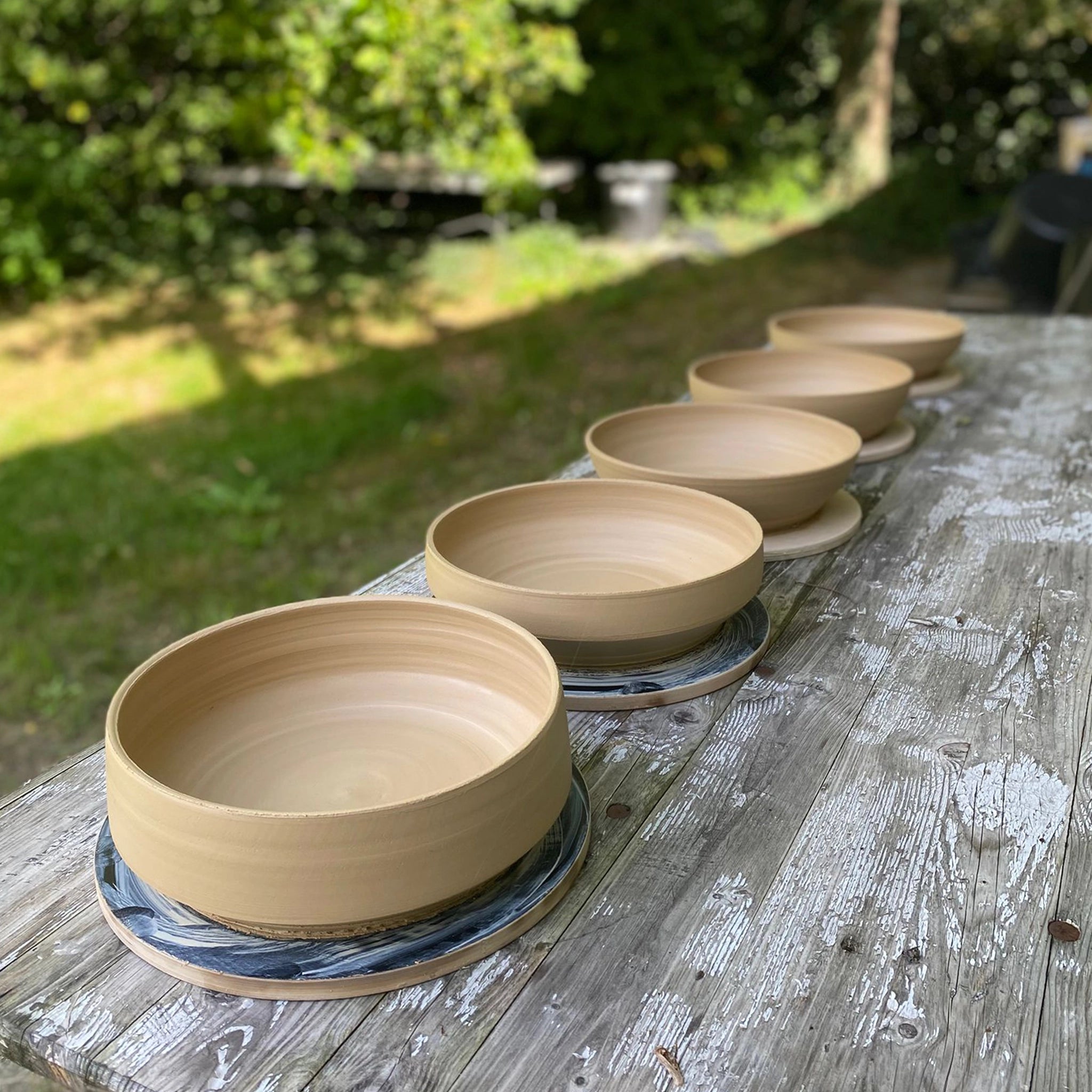 Zes AB Ceramics Serveerschalen Ruw Mint D25 staan op een verweerde houten buitentafel, omringd door groene bladeren en zacht zonlicht.