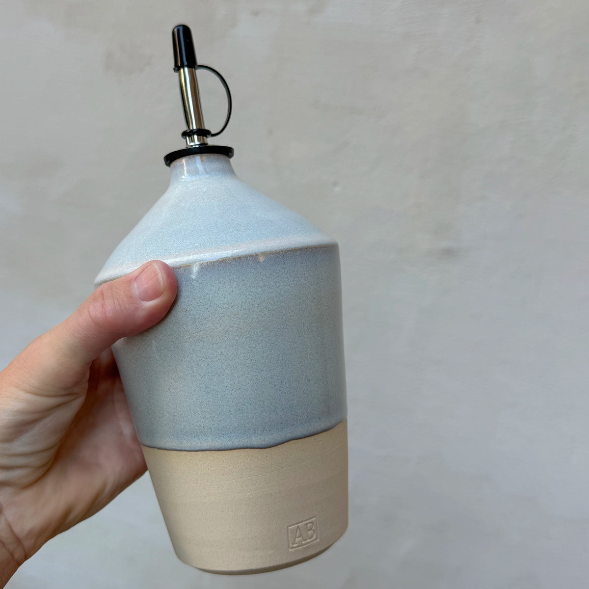 Een hand houdt de AB Ceramics Olijfolie Fles 2 Prai Opaal 70 cl vast tegen een grijze muur, met zicht op de hoekige schouder en de glazuurrand.