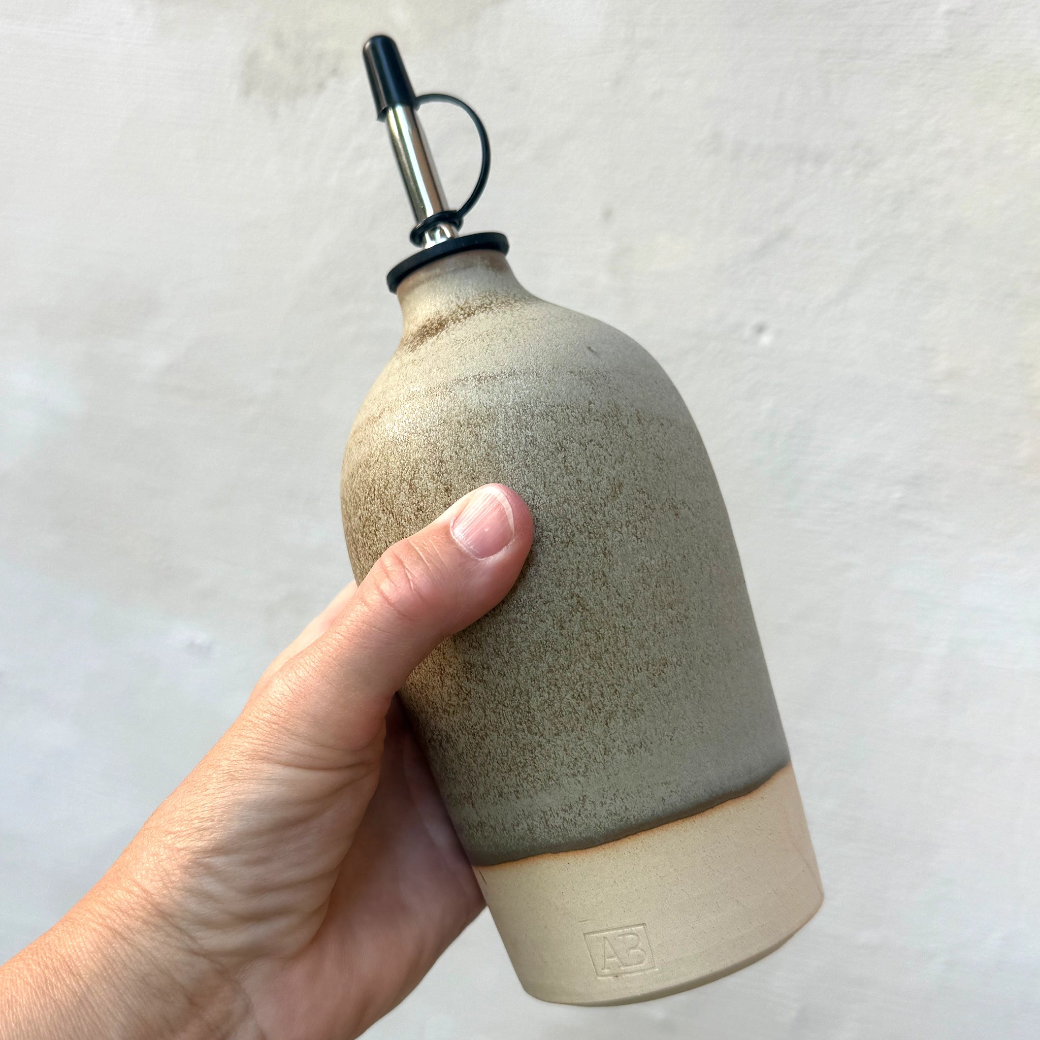 Een hand houdt de AB Ceramics Olijfolie Fles 2 Wit Mos 55 cl vast, een handgeblazen keukenaccessoire van keramiek van AB Ceramics, met een metalen tuit en tweekleurig beige en grijs glazuur voor een natuurlijke, aardse uitstraling.