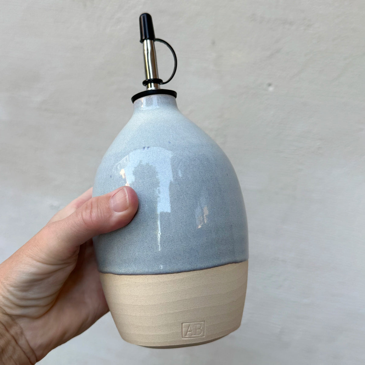 Een hand houdt de eivormige AB Ceramics Olijfolie Fles 4 Prai Opaal 65 cl vast tegen een grijze muur.