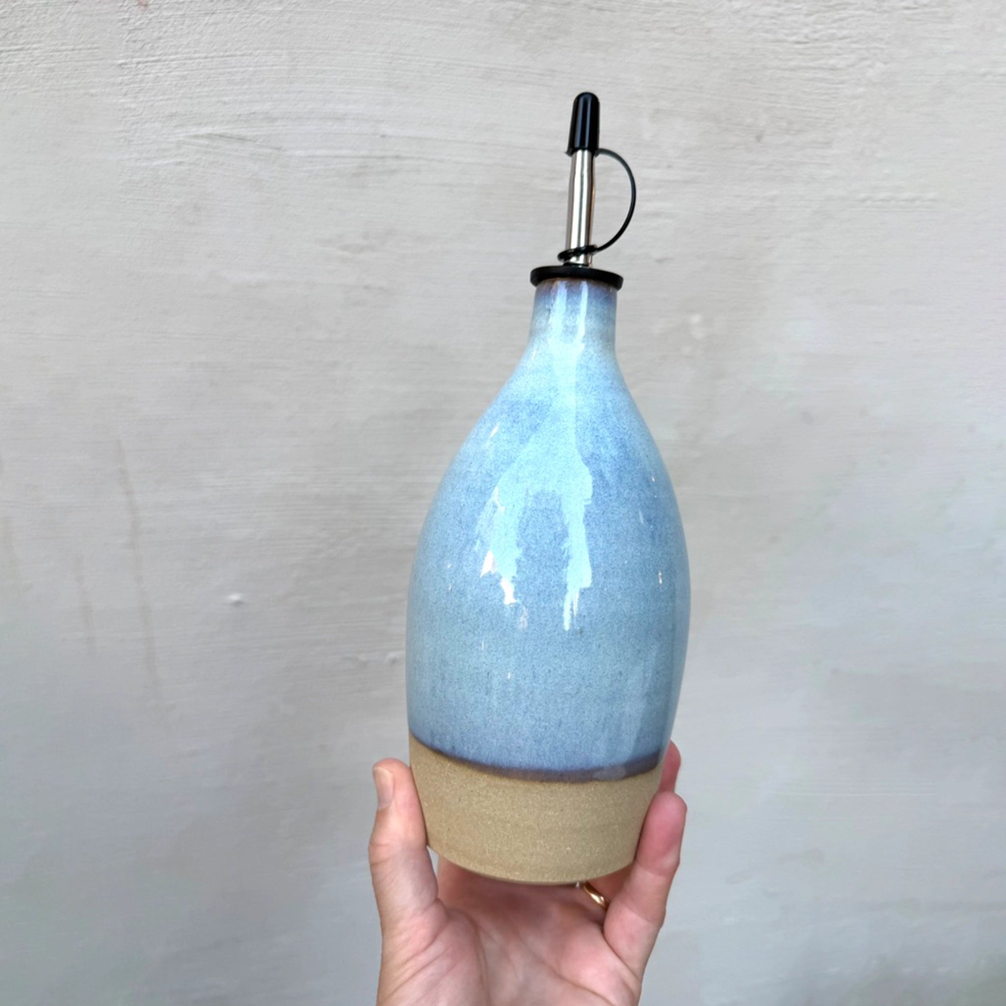 Een hand houdt de AB Ceramics Olijfolie Fles Geel Opaal 61 cl van AB Ceramics vast, met een glanzende lichtblauwe bovenkant, matbeige onderkant en metalen tuit, tegen een effen lichte achtergrond.