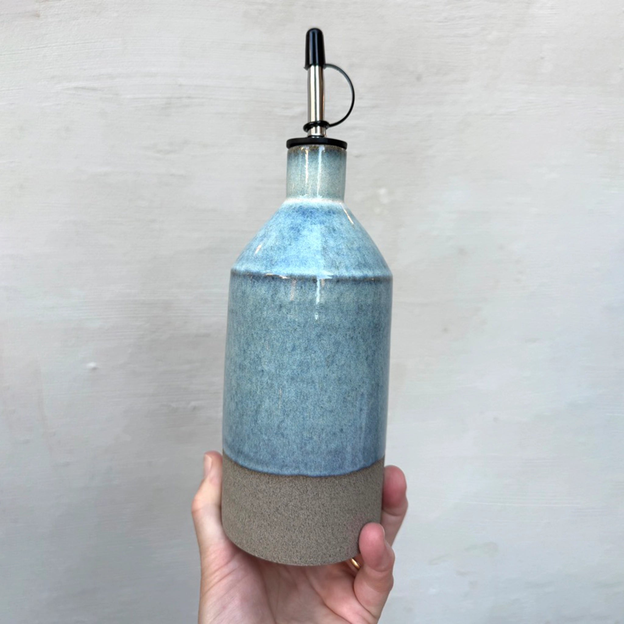 Een hand houdt de AB Ceramics Olijfolie Fles Grijs Opaal 60 cl vast, met opaal glazuur op de lichtblauwe bovenkant, een beige onderkant en een metalen tuit met zwarte dop, tegen een effen lichte achtergrond.