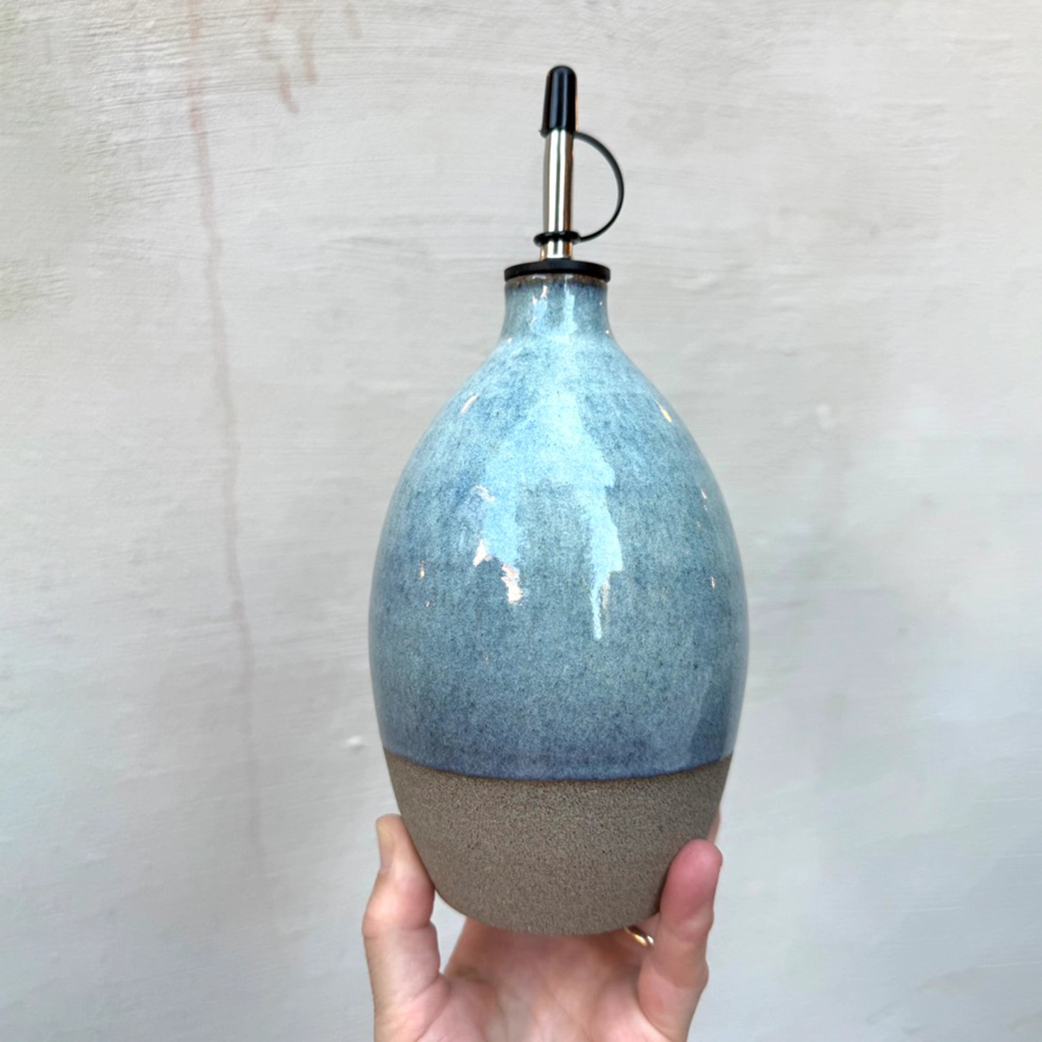 Een hand houdt de AB Ceramics Olijfolie Fles Grijs Opaal 84 cl vast; het handgedraaide keramiek heeft een blauw-grijze glazuur bovenop, een natuurlijke onderzijde en een zwart schenktuitje-stijlvol tegen een lichte, textuurvolle achtergrond.
