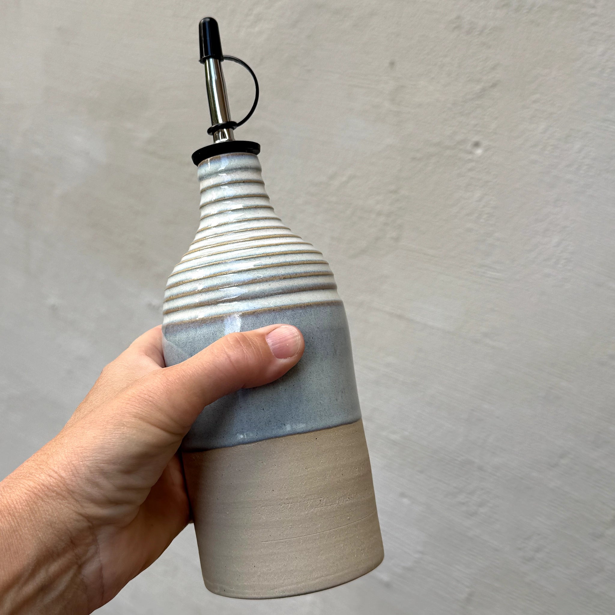 Een hand houdt de AB Ceramics Olijfolie Fles Ribbel Wit Opaal 45 cl vast, met een geribbelde witte opalen bovenkant, gladde onderste helft en een metalen schenktuit, tegen een lichtgrijze wand met textuur.