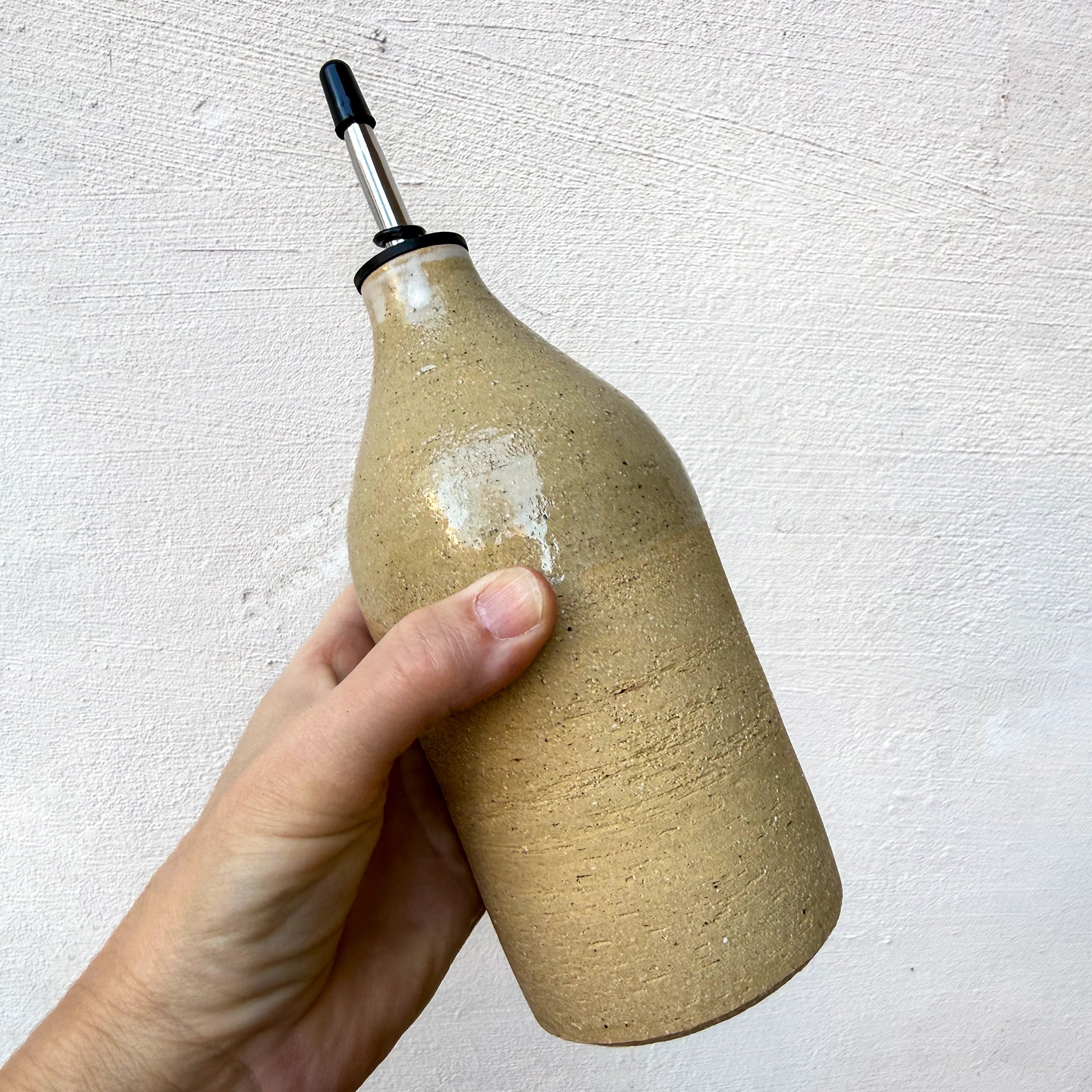 Een hand houdt een AB Ceramics Olijfolie Fles Ruw Transparant 60 cl vast, een beige keramische fles met metalen tuit van AB Ceramics, ideaal voor het schenken van olie of azijn, tegen een lichte achtergrond.
