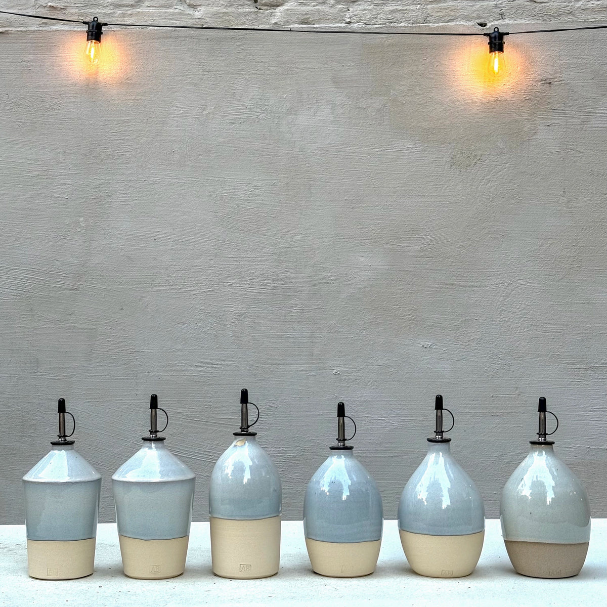 Zes handgedraaide AB Ceramics olijfolieflessen, elk met een andere vorm, op een witte tafel tegen een grijze muur met twee brandende lampjes.