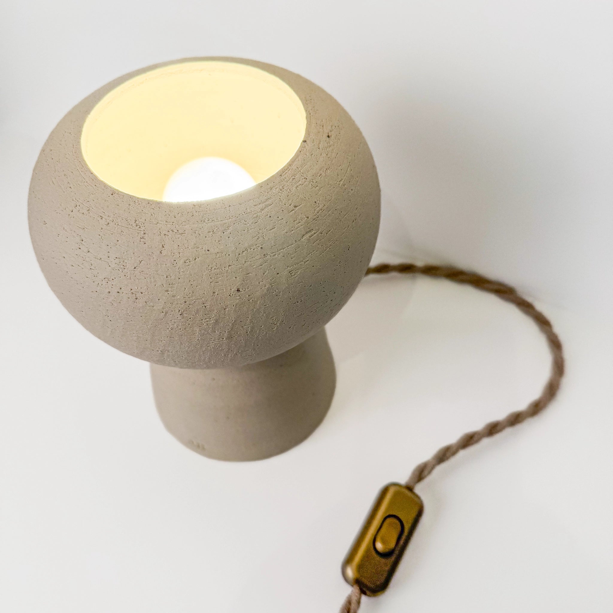 De AB Ceramics Sfeerlamp 20cm van AB Ceramics is een kleine, ronde lamp van beige keramiek met een getextureerde afwerking, gouden aan/uit-schakelaar aan een gevlochten bruin snoer en straalt zacht warm licht uit vanuit de open bovenkant.