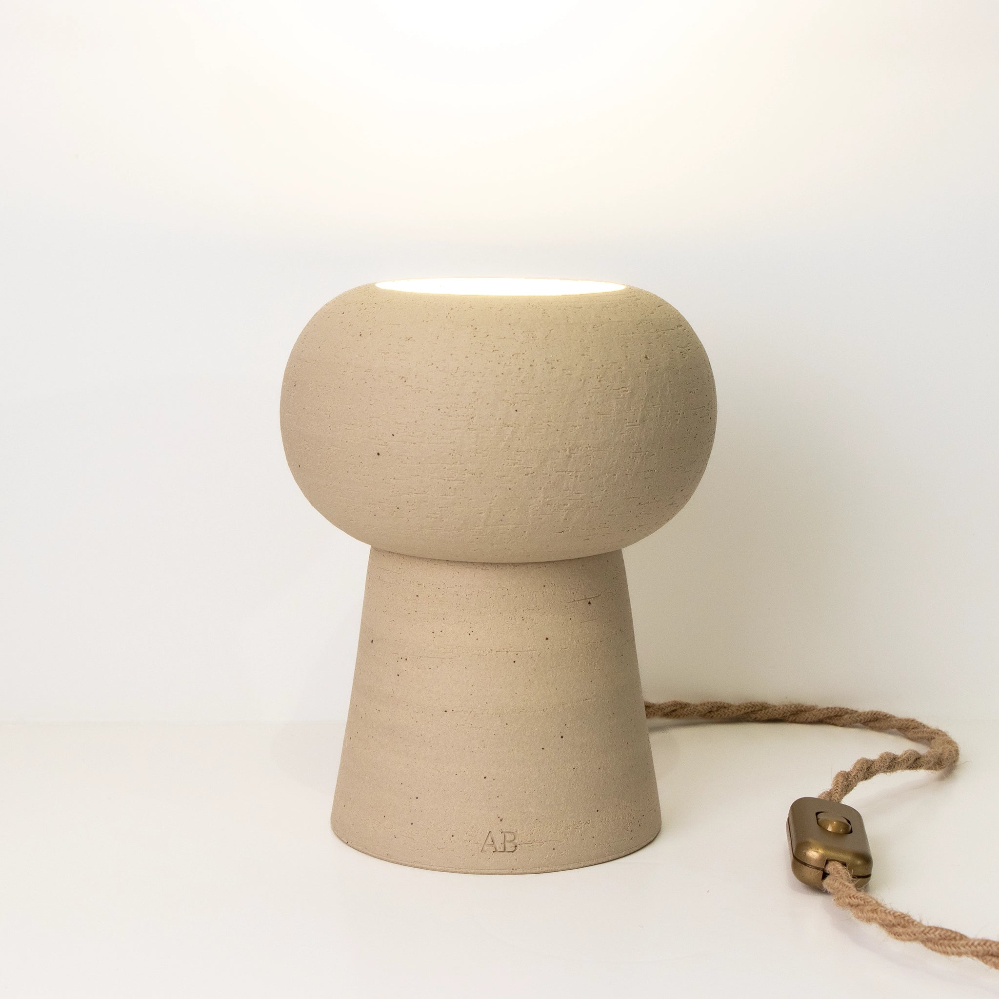 De AB Ceramics Sfeerlamp 20cm is een handgemaakte keramieken lamp in beige met afgeronde bovenkant, conische basis en bruin gevlochten snoer met bronzen schakelaar. Geplaatst op een witte ondergrond, tegen een witte achtergrond.