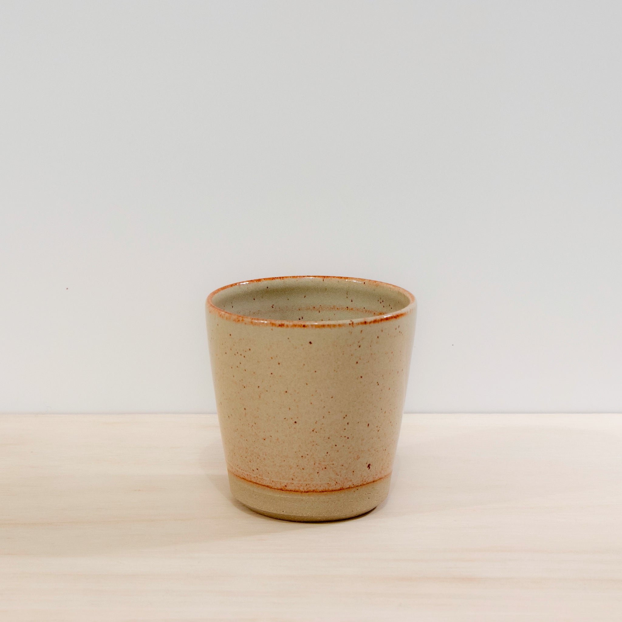 De AB Ceramics Beker Geel Beige 20 cl van AB Ceramics is handgedraaid uit gele steengoedklei, heeft een licht gespikkeld beige oppervlak en staat op een lichte houten ondergrond met een witte achtergrond.