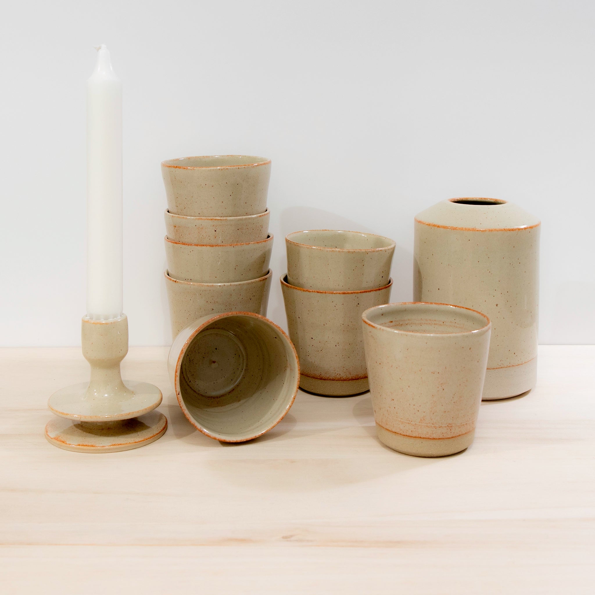 Een selectie serviesgoed van AB Ceramics wordt tentoongesteld op licht hout tegen een witte achtergrond, met de AB Ceramics Beker Geel Beige 20 cl, twee vazen, een kandelaar met witte kaars en een kom van gele steengoedklei.