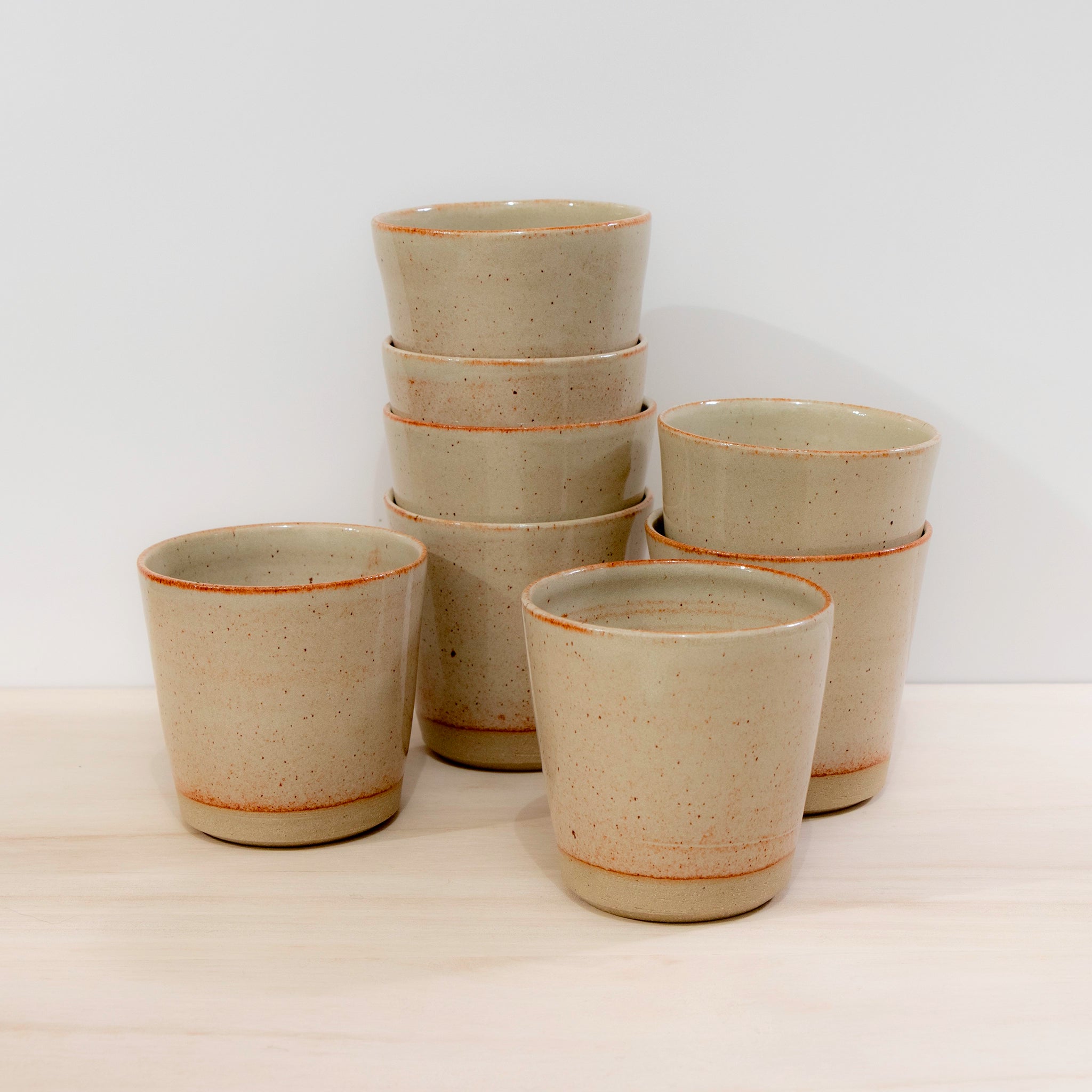 Acht AB Ceramics Beker Geel Beige 20 cl kopjes, handgemaakt van beige steengoed met oranje spikkels, staan op licht hout: vijf gestapeld, drie vooraan, tegen een effen witte achtergrond.
