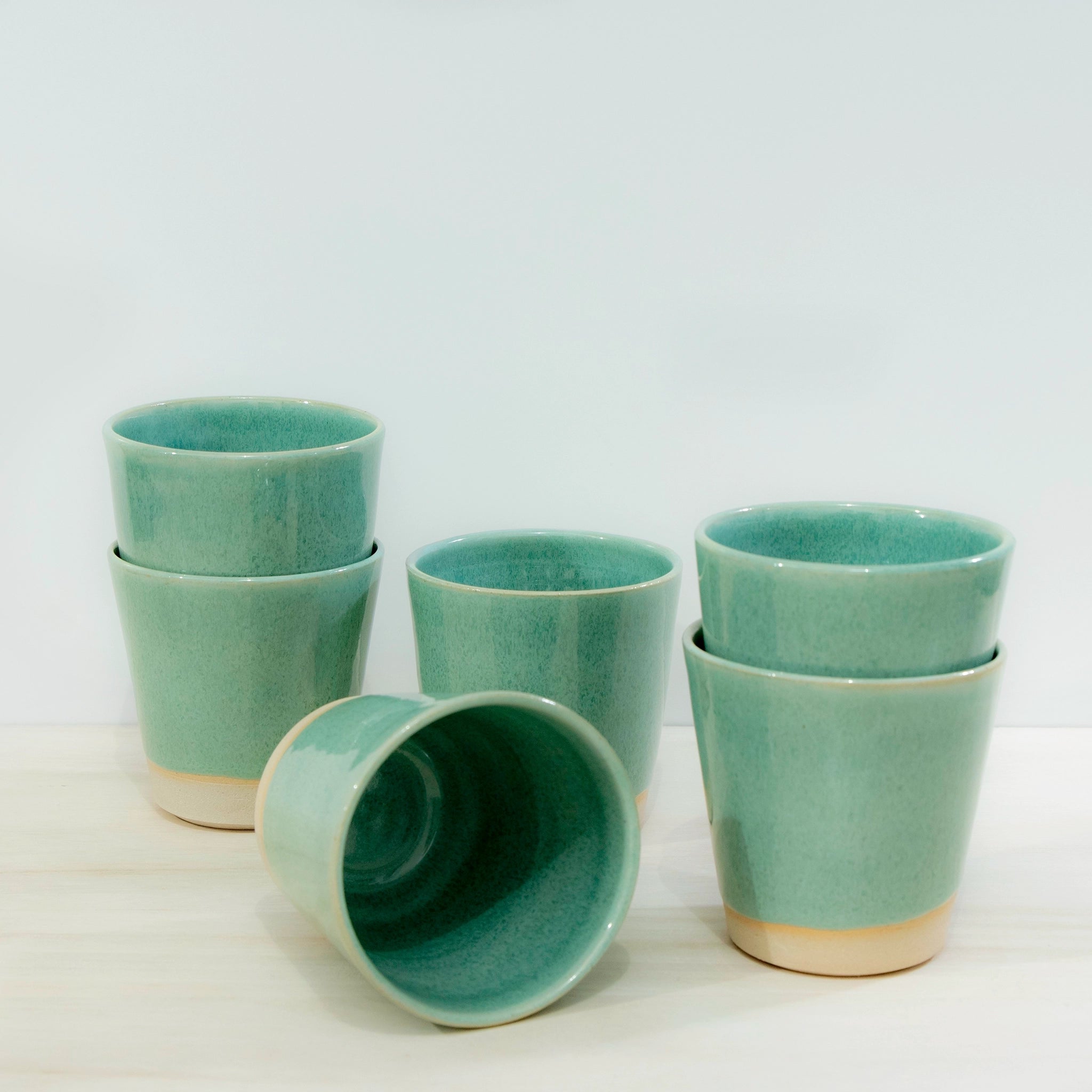 Zes AB Ceramics Beker Wit Groen 20 cl-steengoed kopjes met beige bodems staan op een licht oppervlak - twee gestapeld, een op zijn kant liggend en een rechtop - tegen een lichte achtergrond.