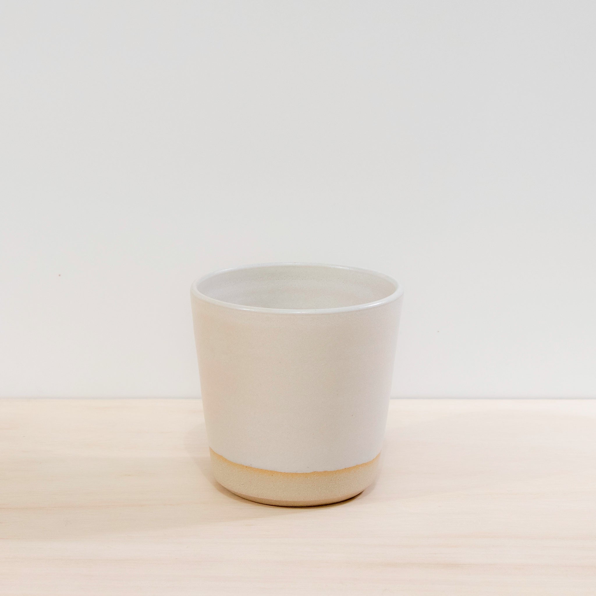 De AB Ceramics Beker Wit Mat Wit 20 cl van AB Ceramics is een handgedraaide beker van witte steengoedklei met een lichtbeige bodem, getoond op een licht houten oppervlak tegen een witte achtergrond.