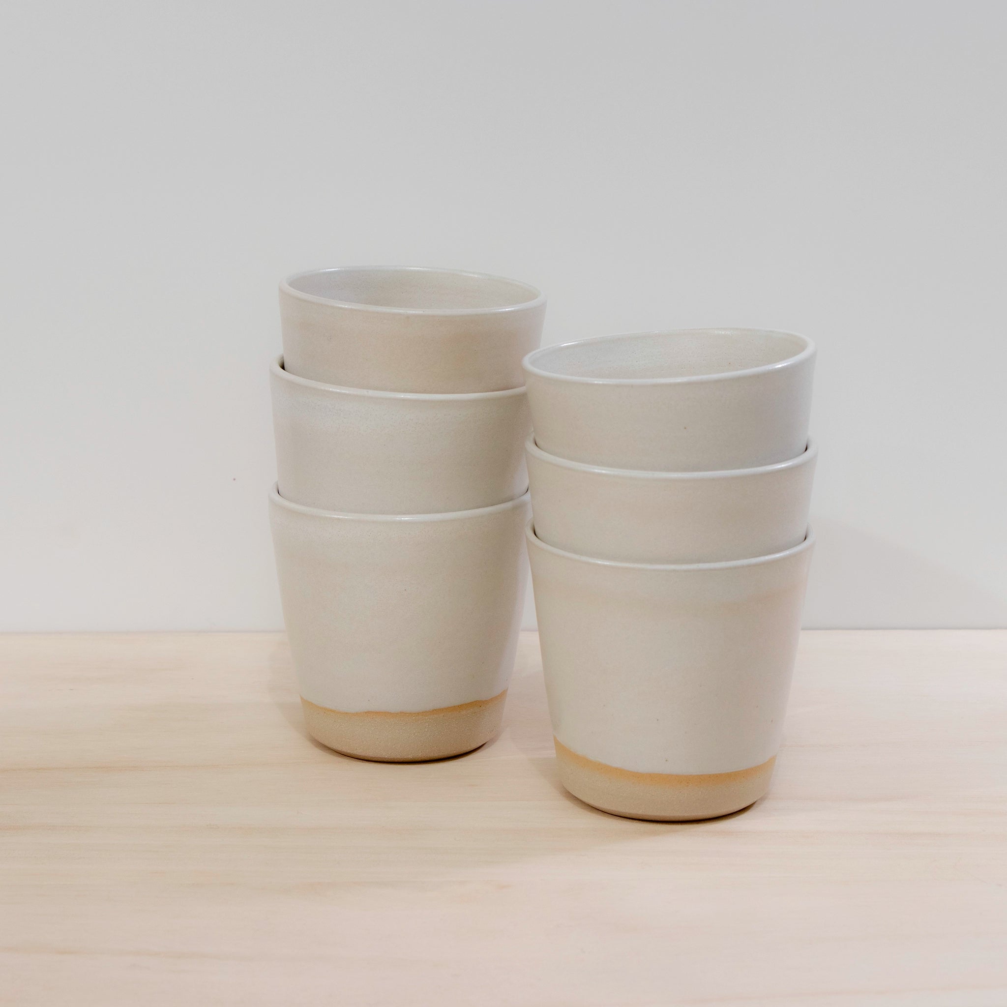 Zes AB Ceramics Beker Wit Mat Wit 20 cl bekers zijn gestapeld in twee kolommen op licht hout, met hun lichtbruine basis zichtbaar tegen een eenvoudige witte achtergrond.
