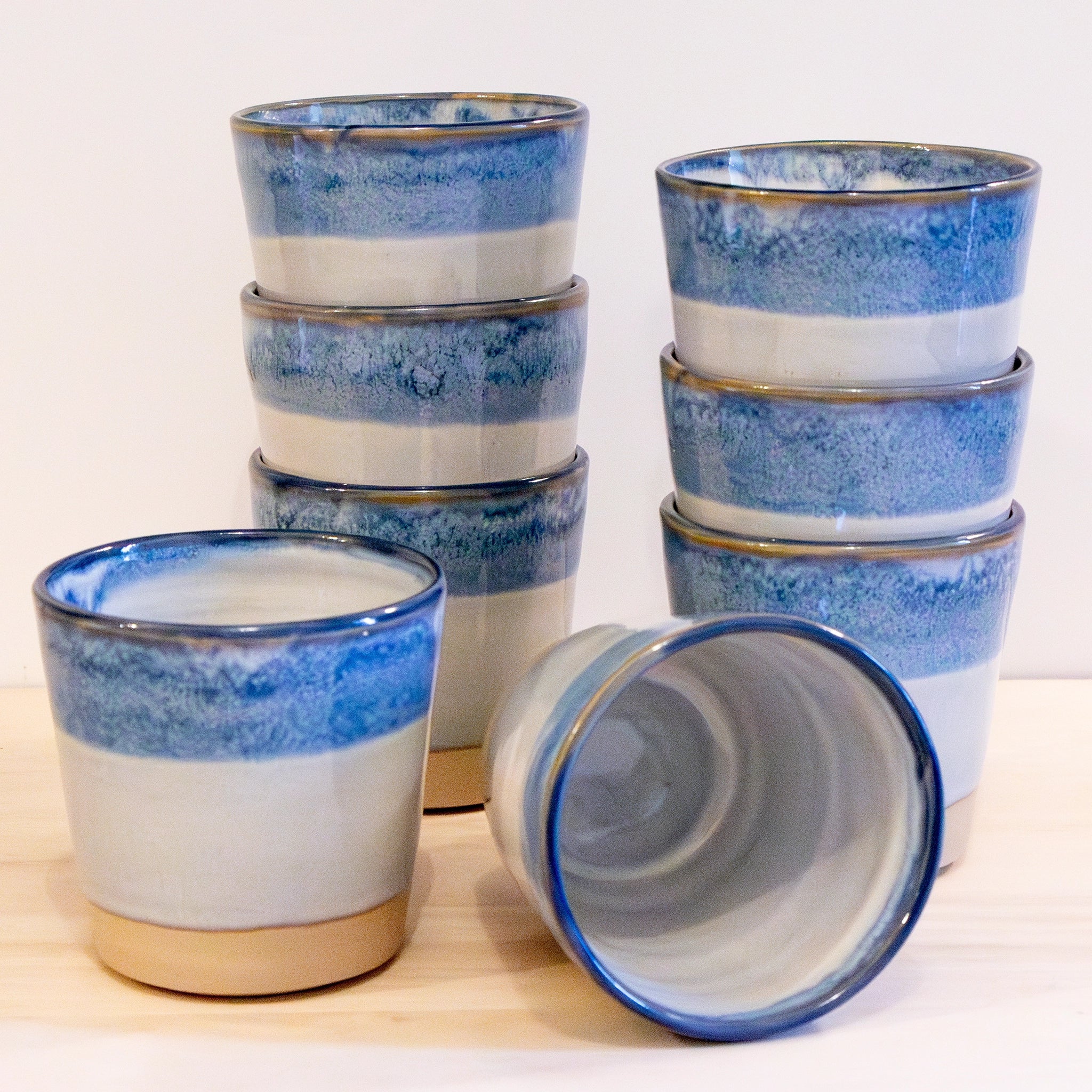 Acht AB Ceramics Beker Wit Opaal/Blauw 20 cl zijn gestapeld in twee ongelijke kolommen, met twee extra unieke exemplaren ervoor, één rechtop en één op zijn zij, op een lichte houten ondergrond.