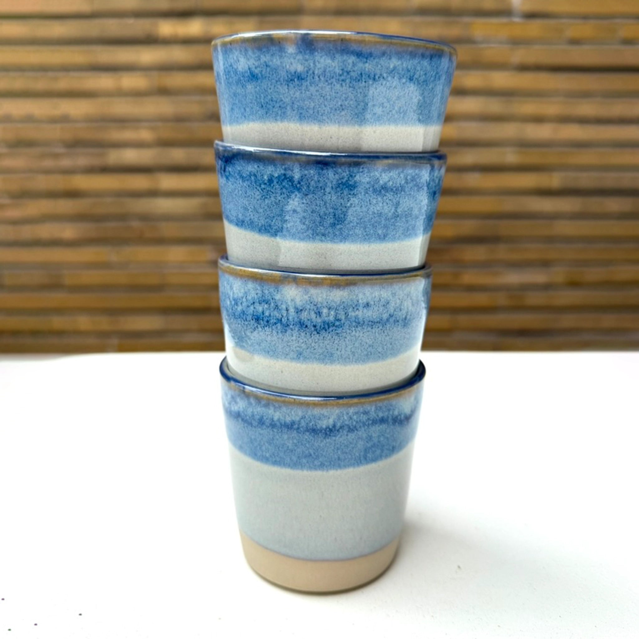 Vier AB Ceramics Beker Wit Opaal/Blauw 20 cl keramische bekers, elk handgemaakt van steengoed klei, zijn gestapeld op een wit oppervlak met een houten lattenachtergrond.