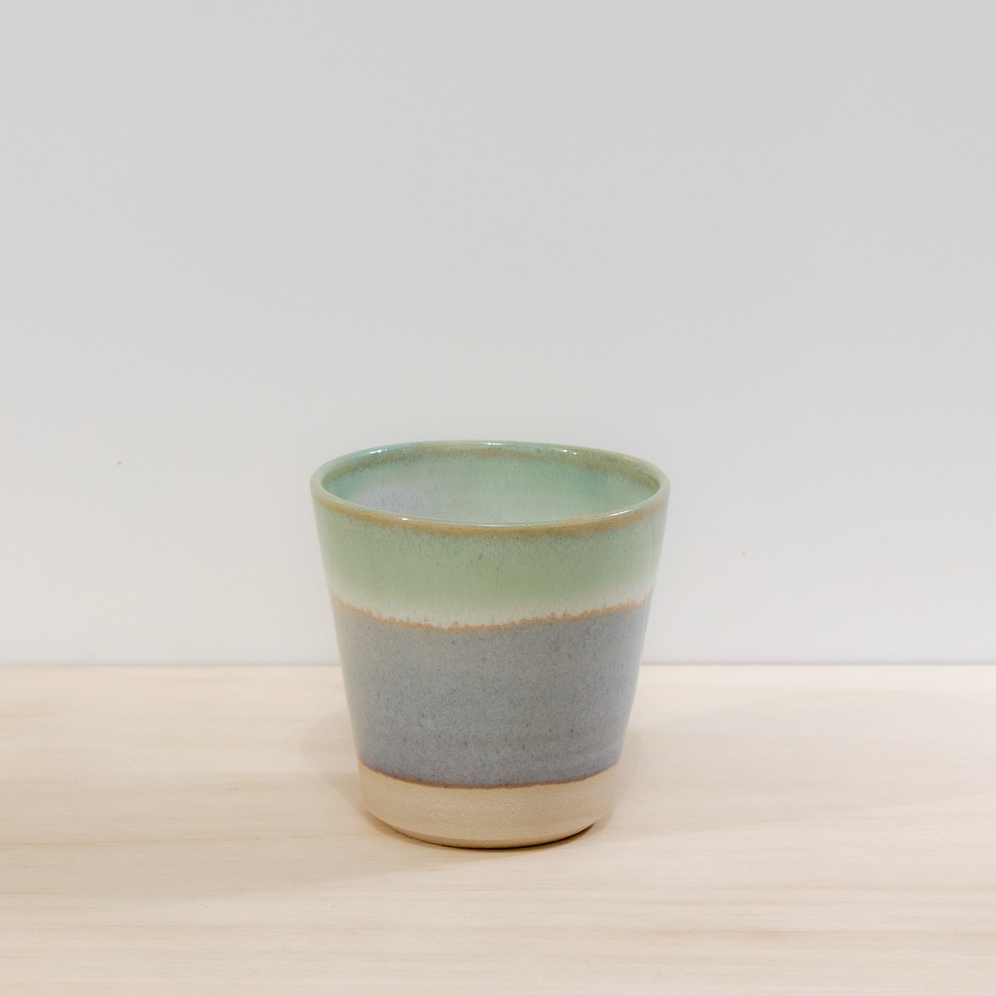 De AB Ceramics Beker Wit Opaal/Lichtgroen 20 cl, een unieke handgemaakte keramische beker van AB Ceramics met een lichtgroene bovenkant, blauwgrijze band en beige basis, rust op een licht houten oppervlak tegen een effen witte achtergrond.