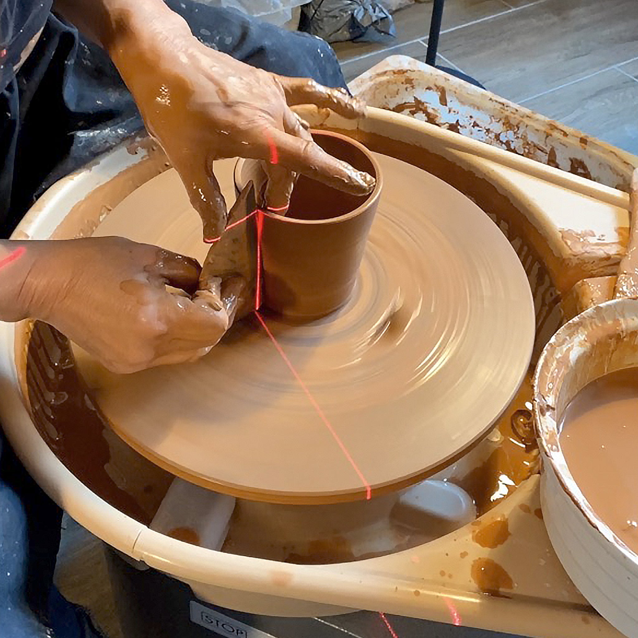Een persoon vormt de AB Ceramics Beker Geel Beige 20 cl van gele steengoed klei met zijn handen en een gereedschap op een spinnewiel, terwijl natte klei en bruine slip het oppervlak bedekken en een rode laserlijn helpt bij het uitlijnen. Merk: AB Ceramics.