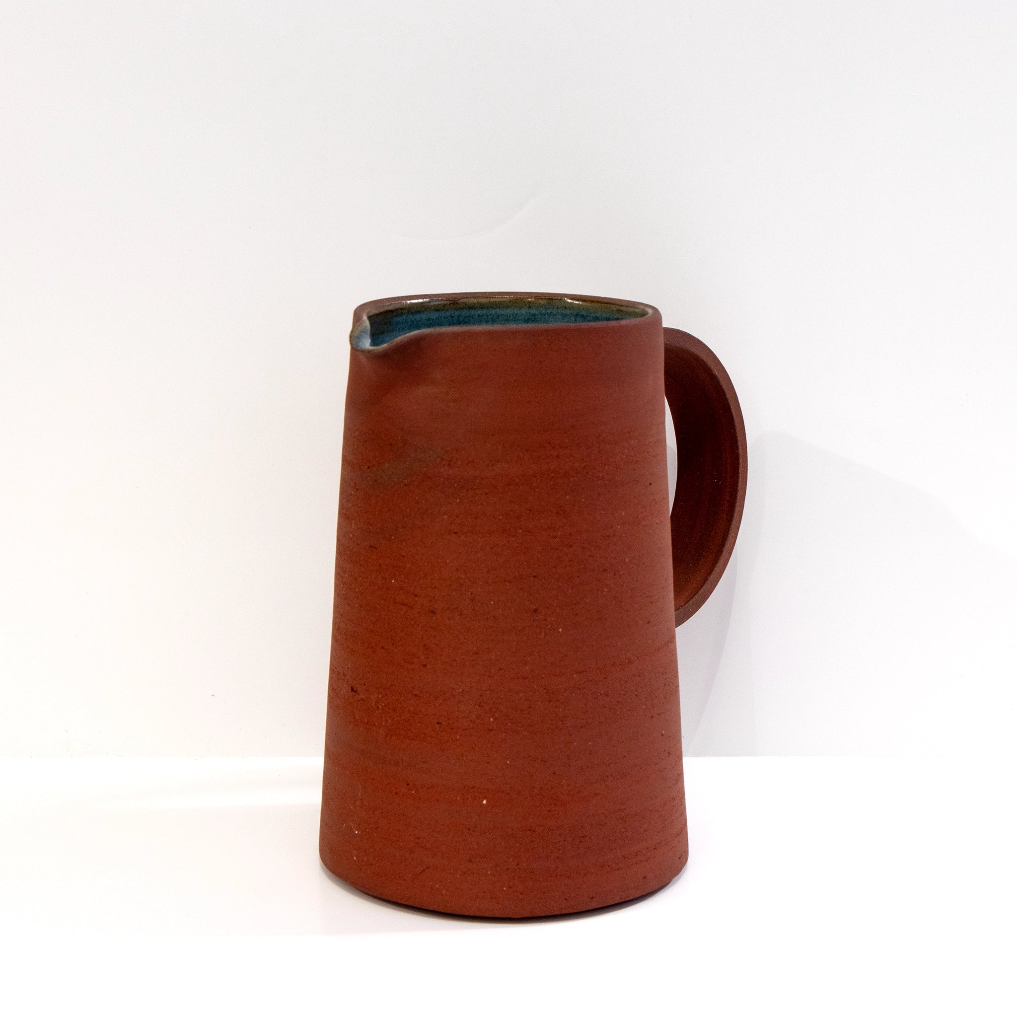 De AB Ceramics Karaf 3 Rood Opaal Blauw 110 cl is een hoge, handgegoten roodbruine karaf van rode klei, met een breed handvat, schenktuit en een blauwgroene binnenkant van opaalglazuur, afgebeeld tegen een effen witte achtergrond.