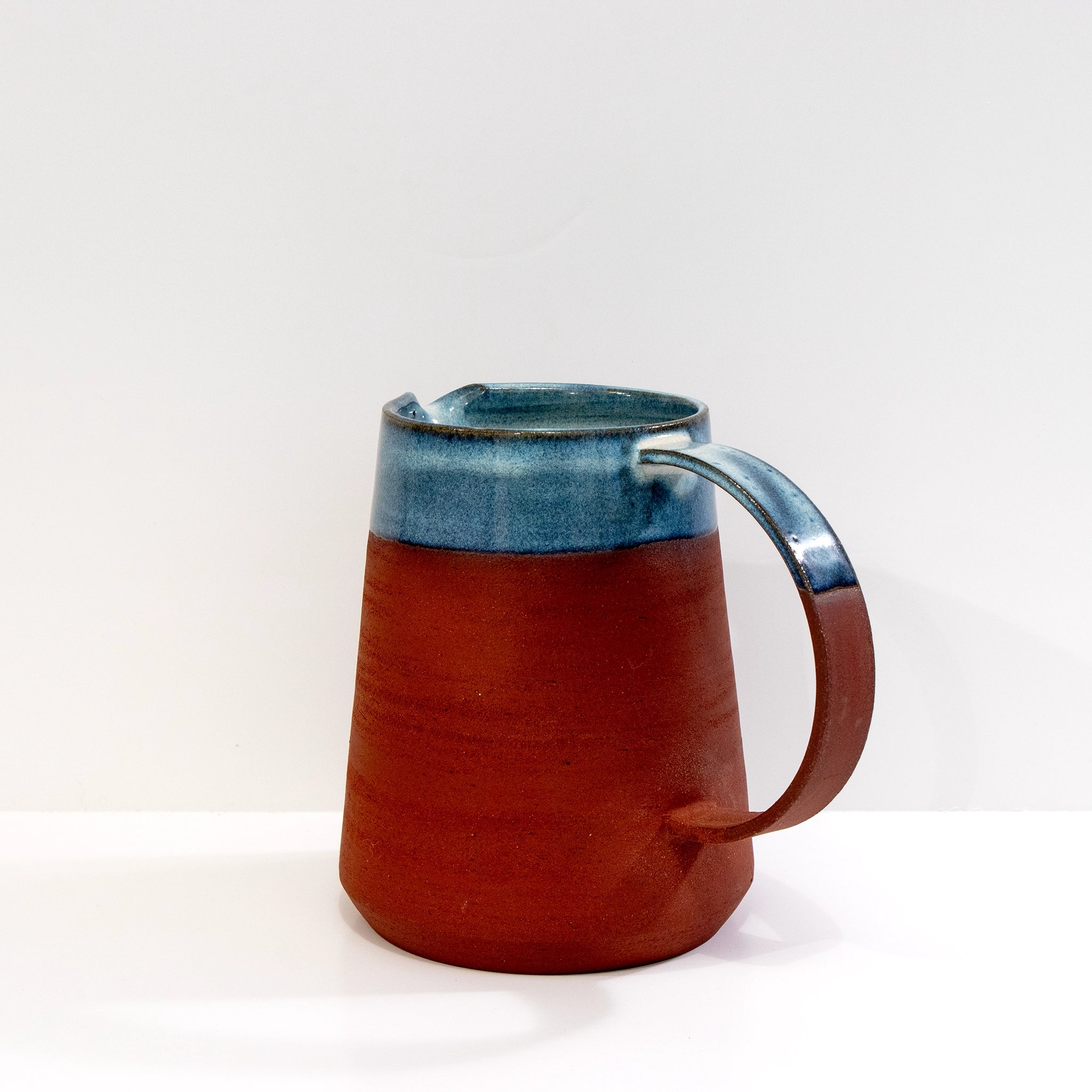 De AB Ceramics Karaf 5 Rood Opaal Blauw 100 cl heeft een handgegoten body van rode klei, een brede basis en opaalglazuur op de bovenste helft en het handvat, tegen een effen witte achtergrond.