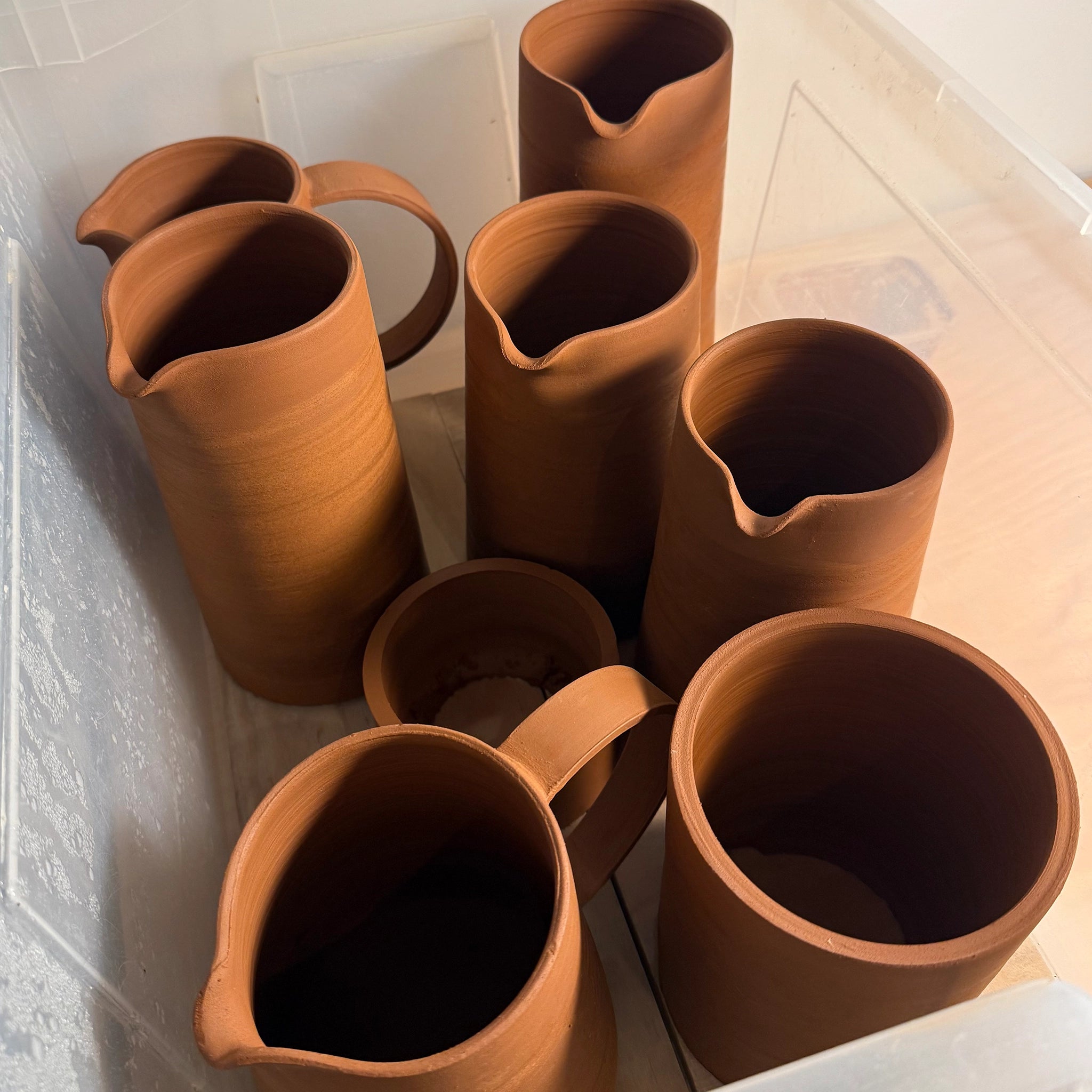 Zes AB Ceramics Karaf 6 Rood Opaal Blauw 50 cl flessen zijn gerangschikt in een plastic container. Ze hebben elk een schenktuit en een handvat. De roodblauwe tinten en gladde afwerking worden benadrukt door het zachte zonlicht.