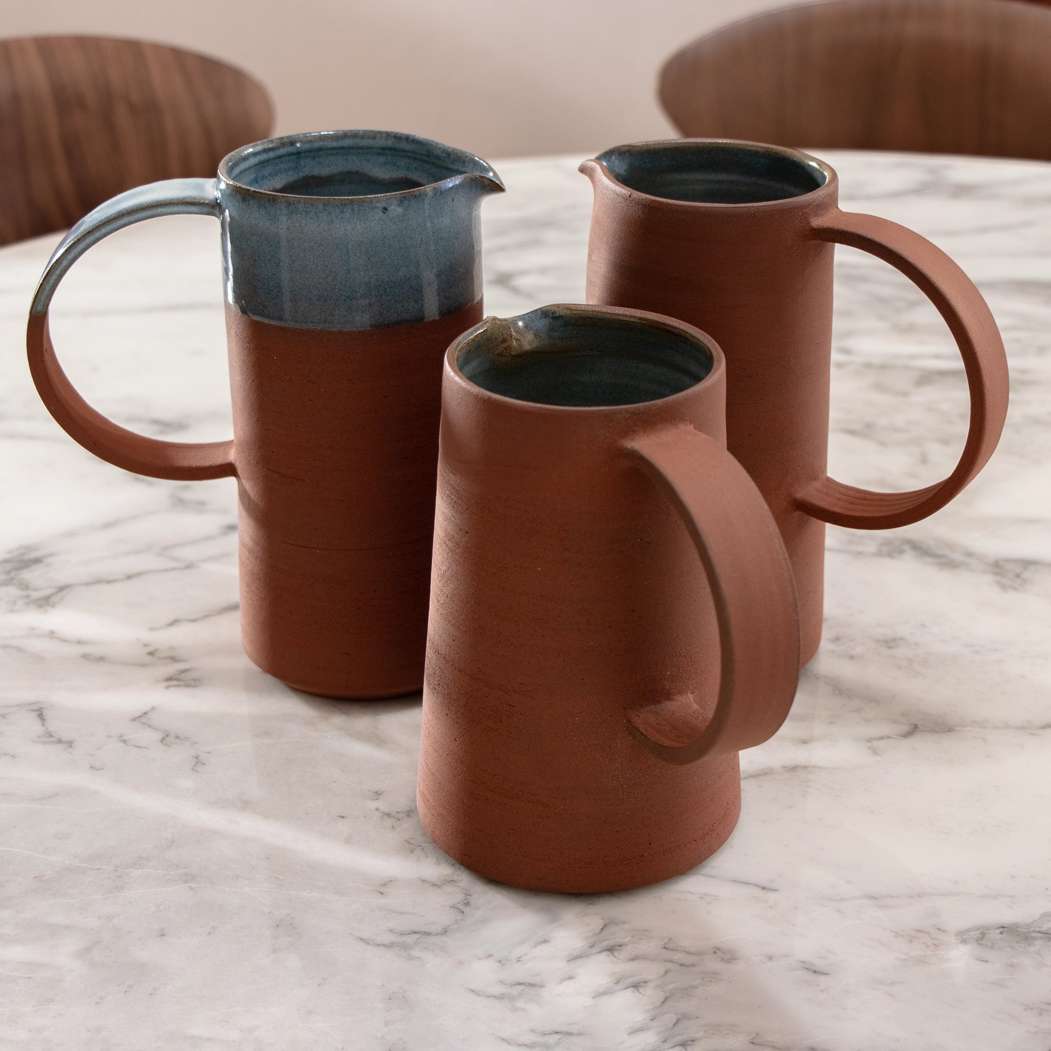 Drie AB Ceramics Karaf 3 Rood Opaal Blauw 110 cl kruiken met grote handvatten, matte afwerking en handgedraaide motieven in bruin rode klei staan op een wit marmeren tafel. Eén heeft opaal glazuur op de rand en aan de binnenkant, met houten stoelen erachter.