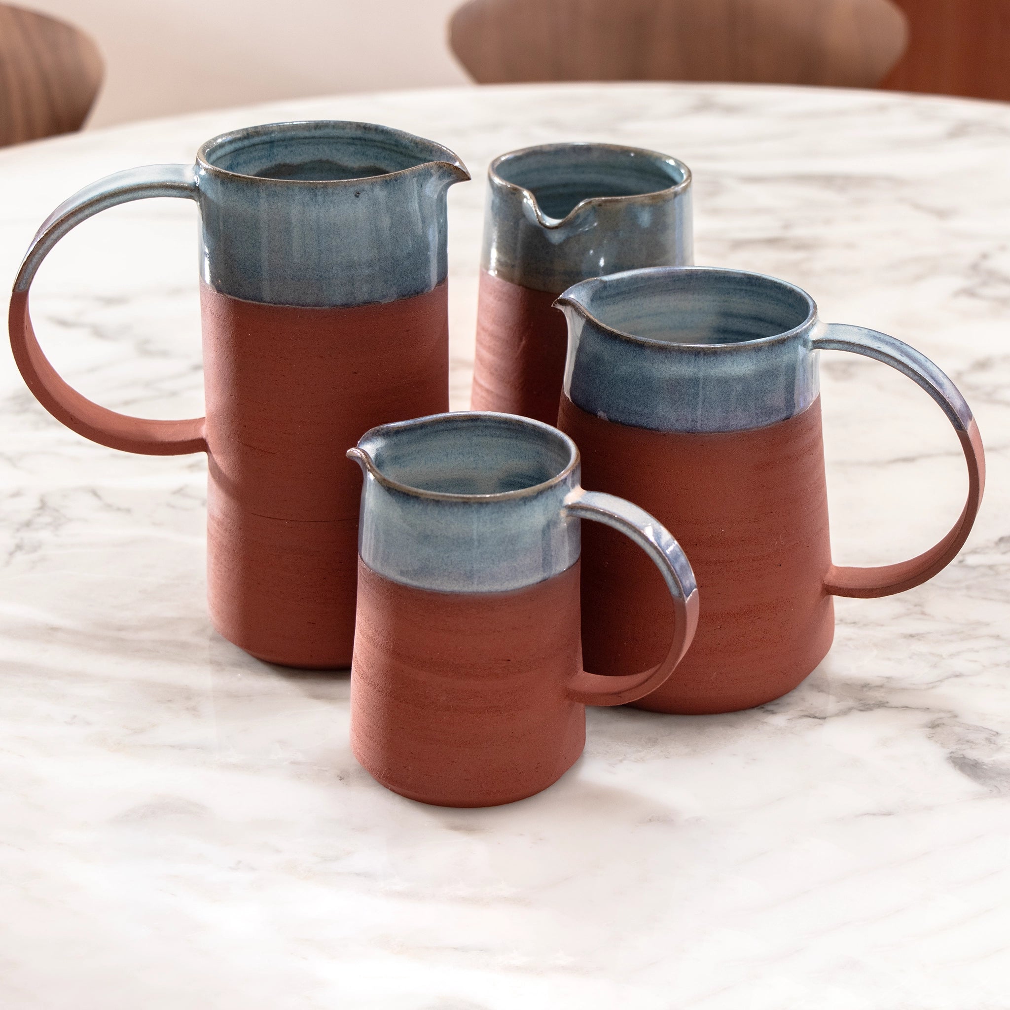 Vier AB Ceramics Karaf 6 Rood Opaal Blauw 50 cl kruiken met grote handgrepen en blauw opaalglazuur staan bij elkaar op een ronde tafel van wit marmer.