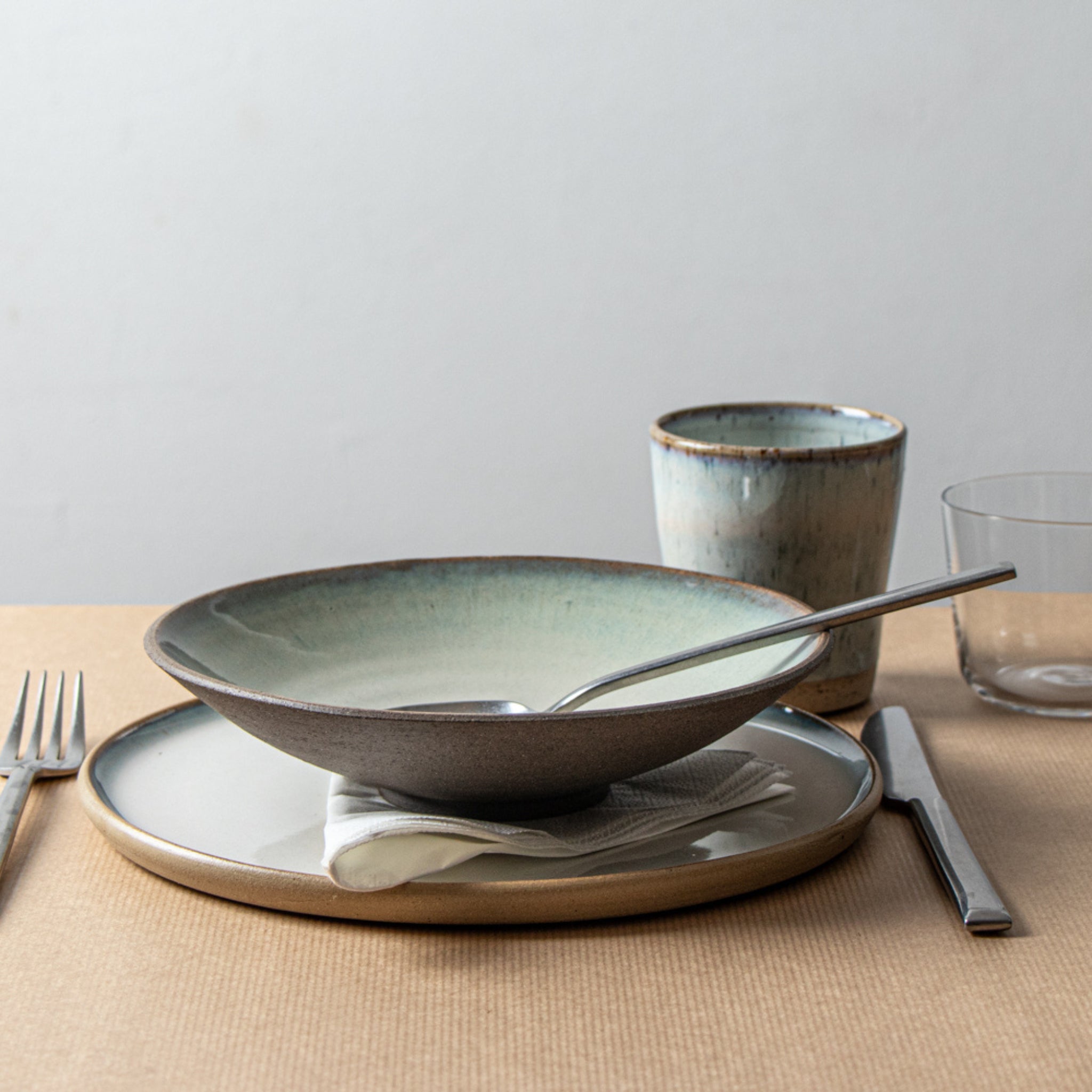 De AB Ceramics Diep Bord Grijs Opaal D21 van AB Ceramics, gemaakt van steengoed klei, wordt getoond met een lepel, gevouwen servet, vork, mes, glas en handgemaakte keramische beker op een beige placemat tegen een effen achtergrond.