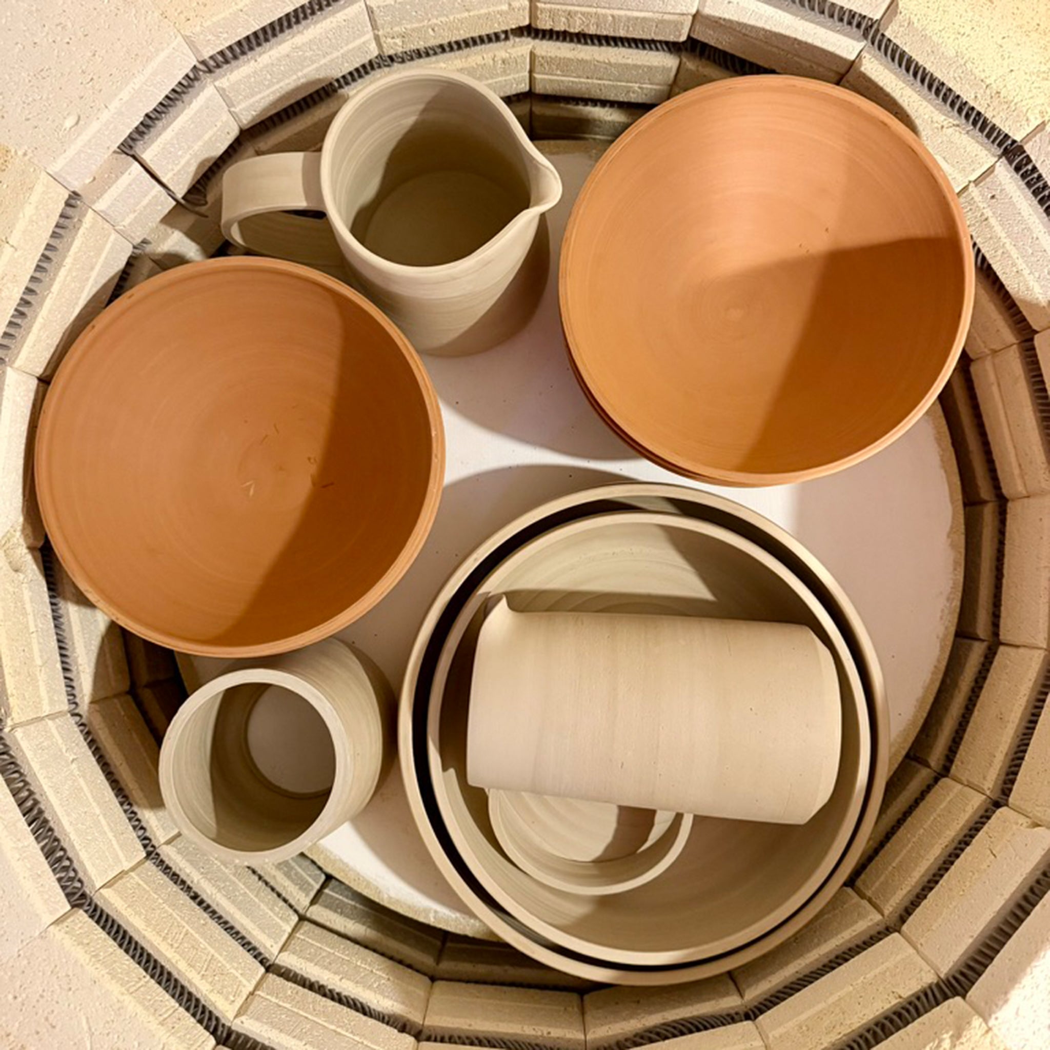 Een AB Ceramics Kom Rood Opaal D18 en andere ongeglazuurde stukken rusten in een ronde oven. Van bovenaf gezien tonen hun roodbruine en beige tinten het kleurengamma van AB Ceramics klei vóór het bakken.