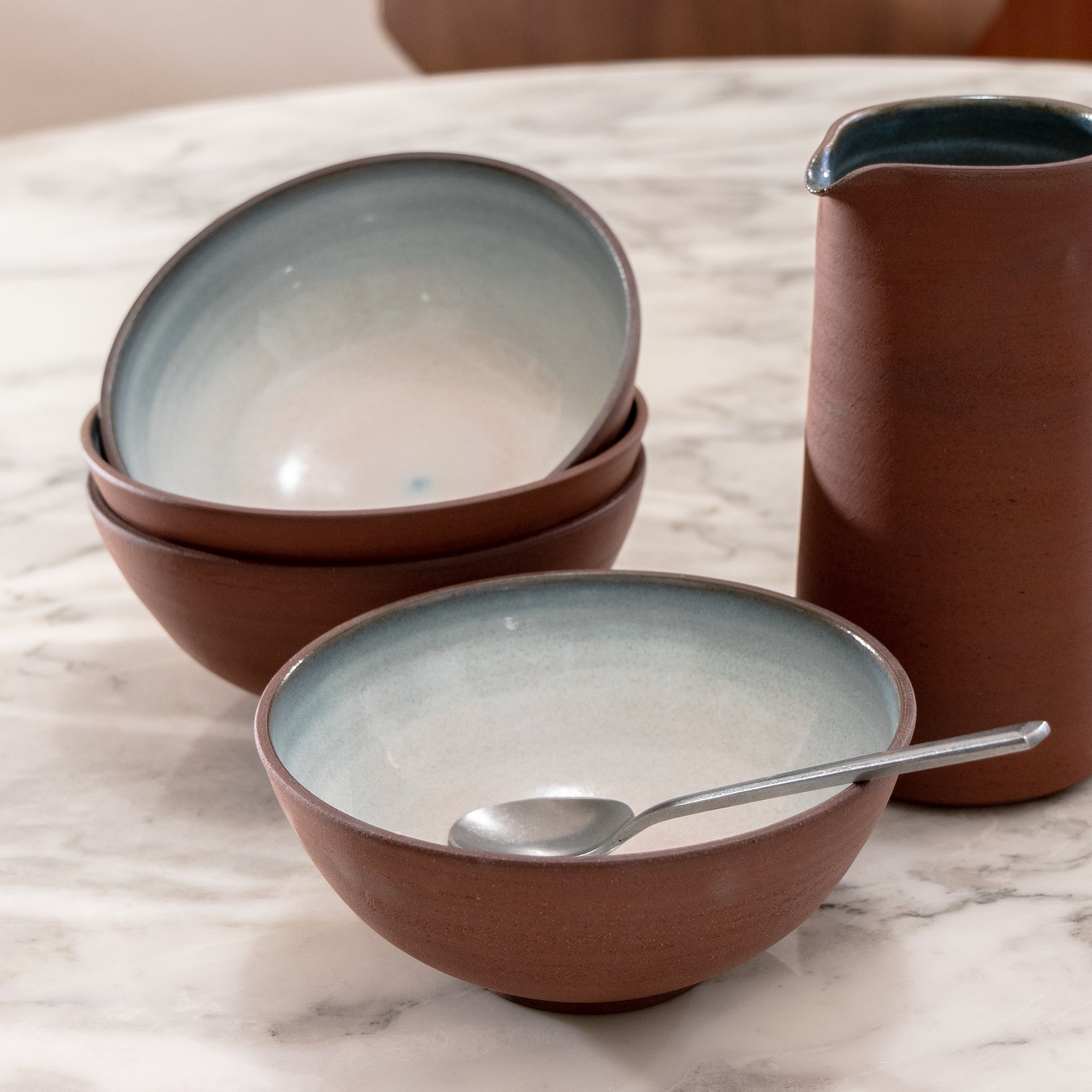 Drie gestapelde AB Ceramics Kom Rood Opaal D18 kommen en een bijpassende noedelkom met lepel, allemaal matbruin van buiten en lichtblauw van binnen, staan op een witmarmeren tafel naast een keramische kan van AB Ceramics.