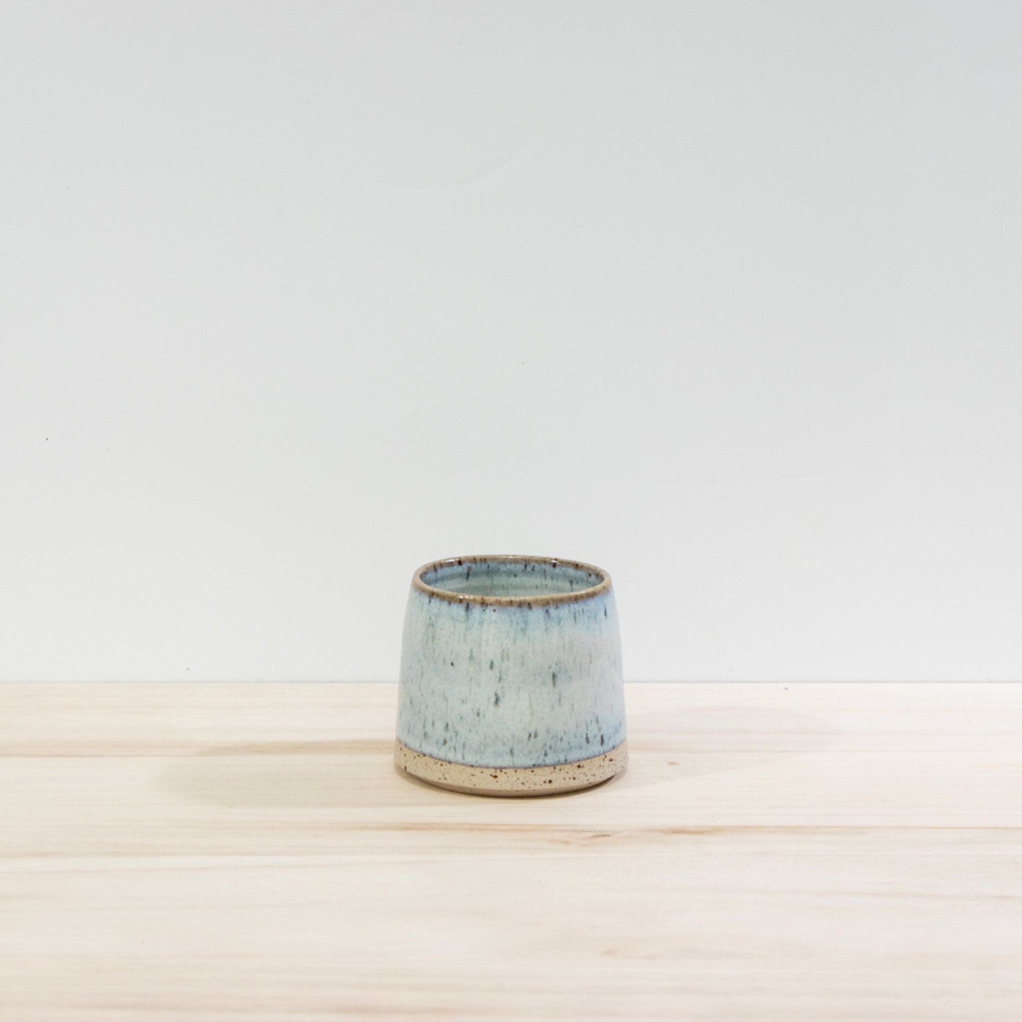 De AB Ceramics Kop Spikkel Opaal D6 is een handgedraaide steengoed kop met lichtblauwe glazuur en subtiele spikkels, geplaatst op een lichte houten ondergrond tegen een witte achtergrond-een uniek ontwerp van AB Ceramics.