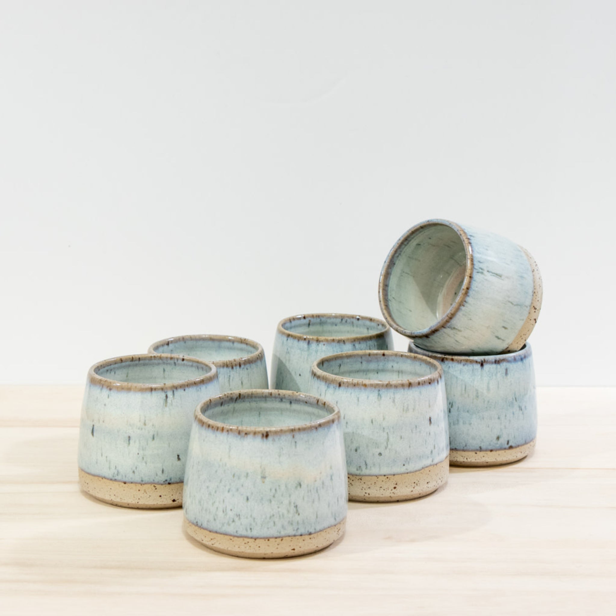 Zes AB Ceramics Kop Spikkel Opaal D6 steengoed koppen met blauw-beige glazuur zijn op licht hout gerangschikt; vijf rechtop, één met uniek design ligt op z'n zij bovenop twee andere koppen.