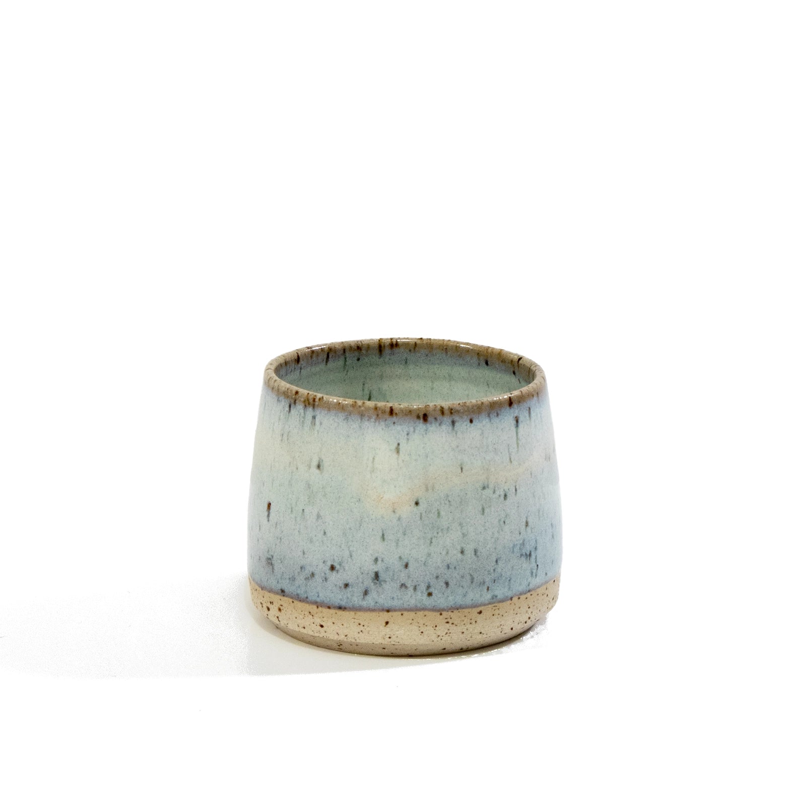 De AB Ceramics Kop Spikkel Opaal D6 is een kleine, ronde steengoed beker met een wijde opening, lichtblauw opaalglazuur en bruine spikkels op een natuurlijk beige basis, handgemaakt door AB Ceramics en afgebeeld op een witte achtergrond.