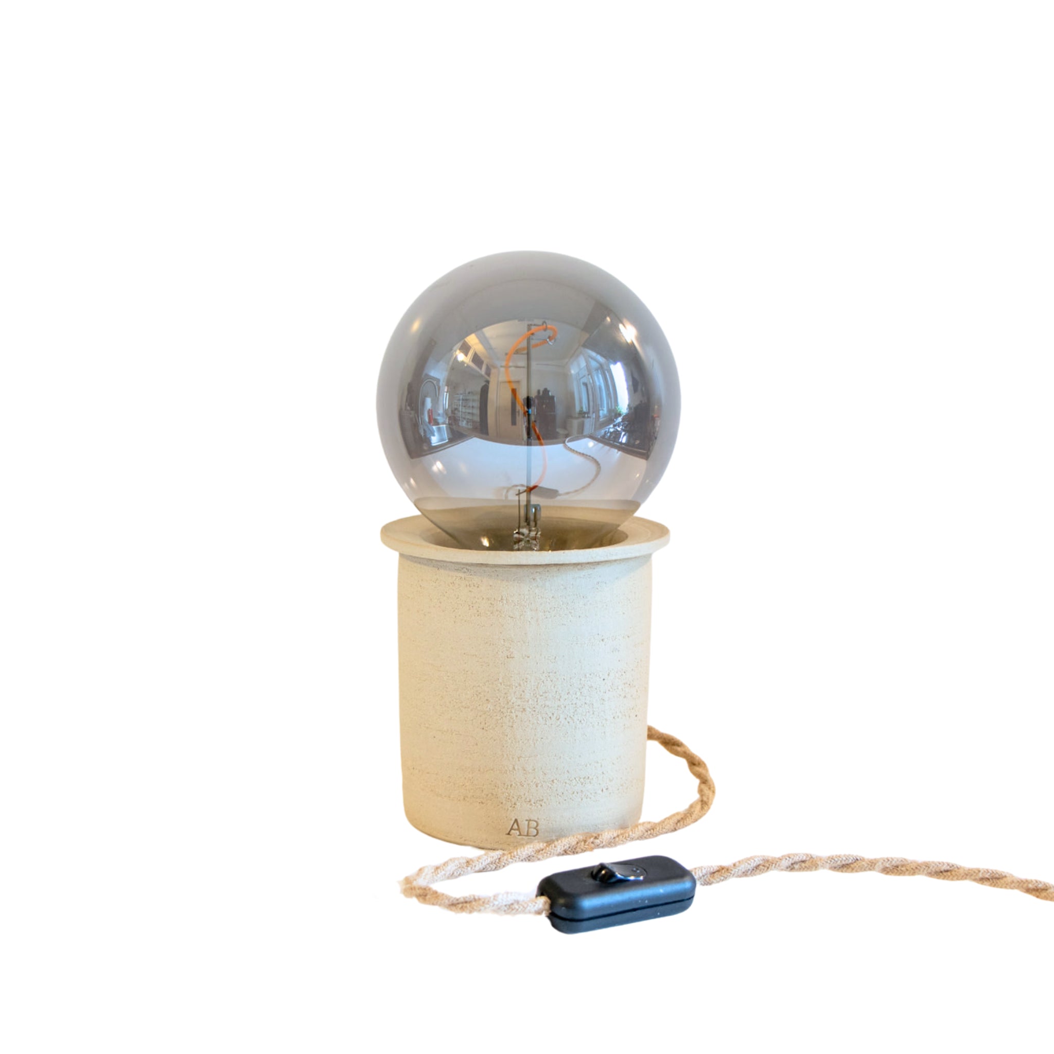 De AB Ceramics LED Lamp van AB Ceramics heeft een ronde, transparante lamp met zichtbare gloeidraad op een cilindrische, licht keramische voet en is inclusief een gevlochten taankleurig snoer met een zwarte schakelaar. De handgemaakte lamp steekt af tegen een witte achtergrond.