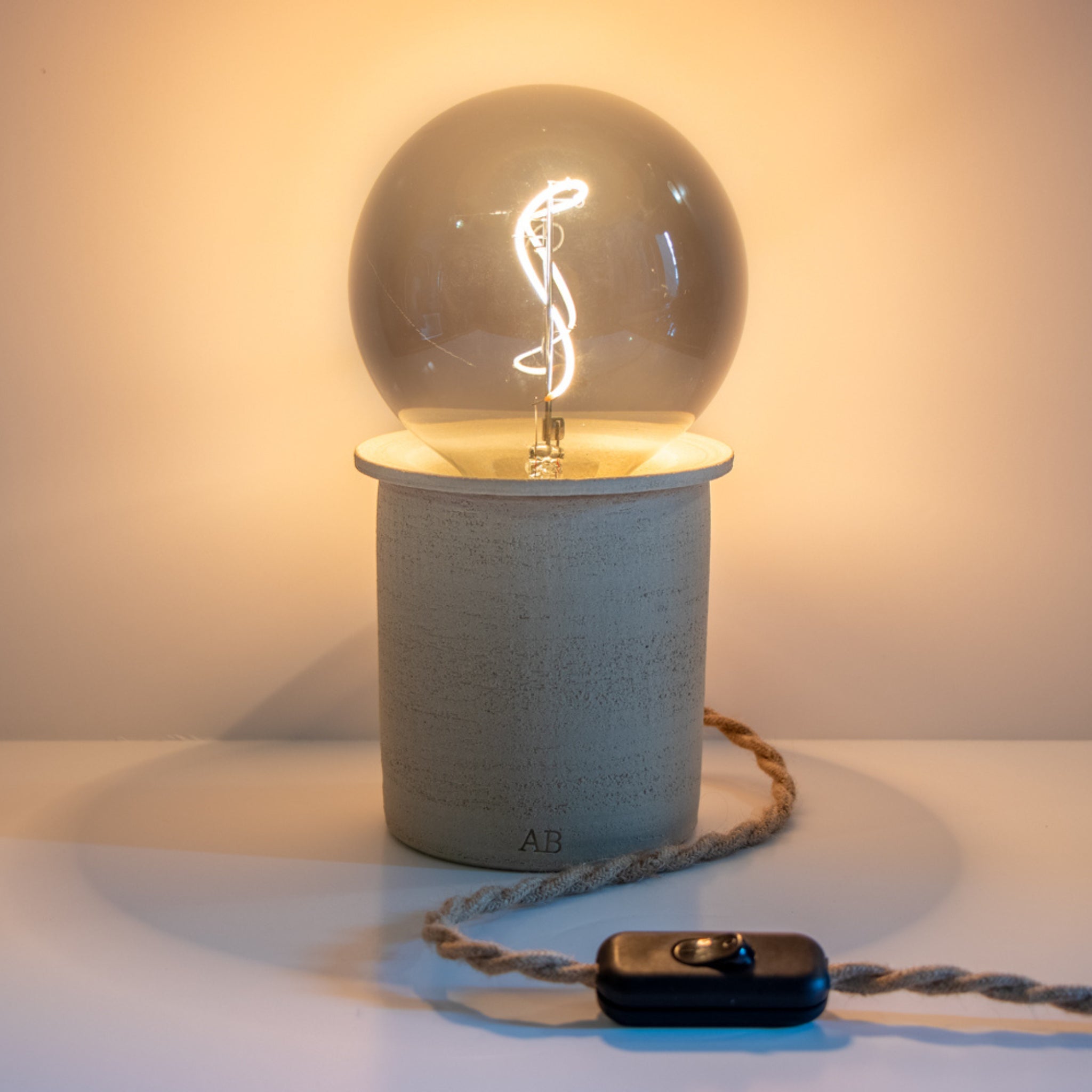 De AB Ceramics LED Lamp van AB Ceramics heeft een ronde gloeilamp op een beige keramische voet. Een gedraaid koord is verbonden met een zwarte schakelaar op een wit oppervlak en werpt warm licht op de achtergrond.