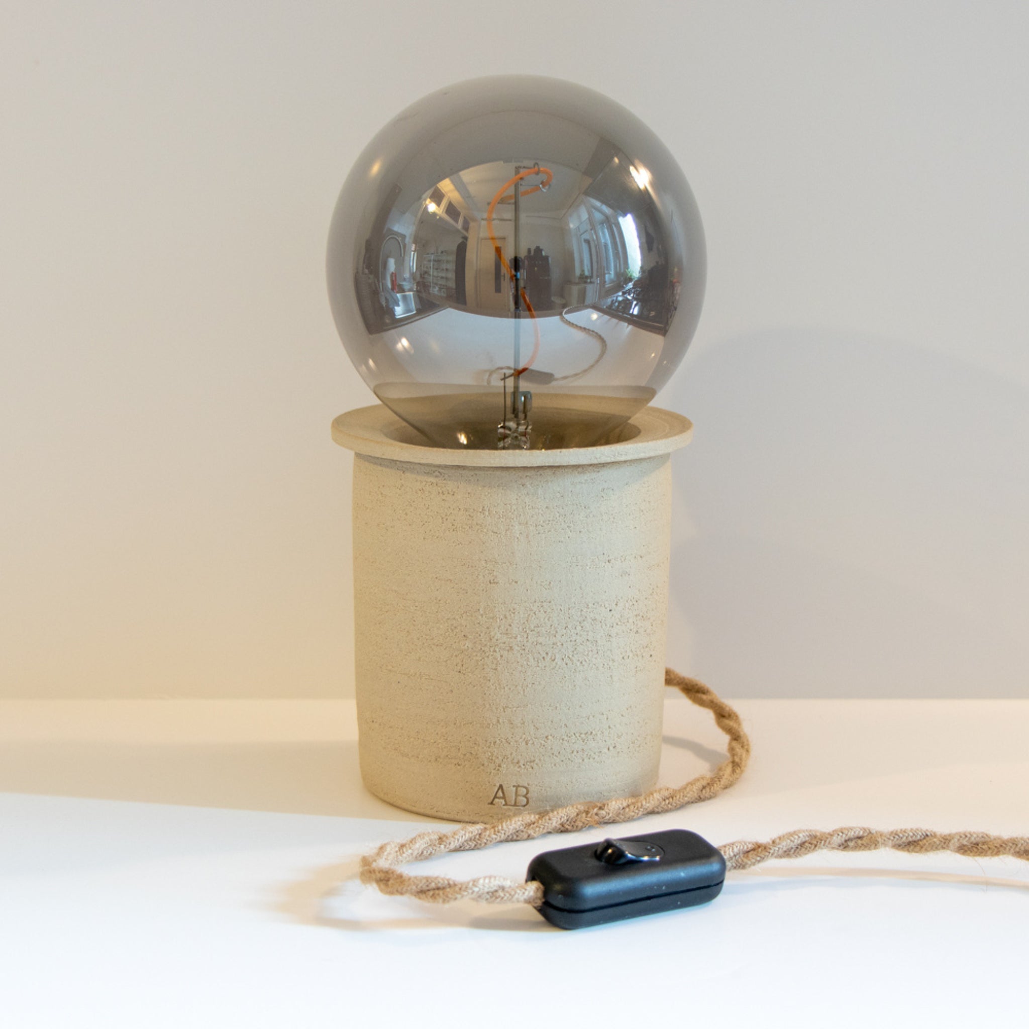 De AB Ceramics LED Lamp heeft een ronde, reflecterende lamp op een beige keramiek voet. Dit handgemaakte stuk van AB Ceramics heeft een gevlochten netsnoer en een zwarte schakelaar.