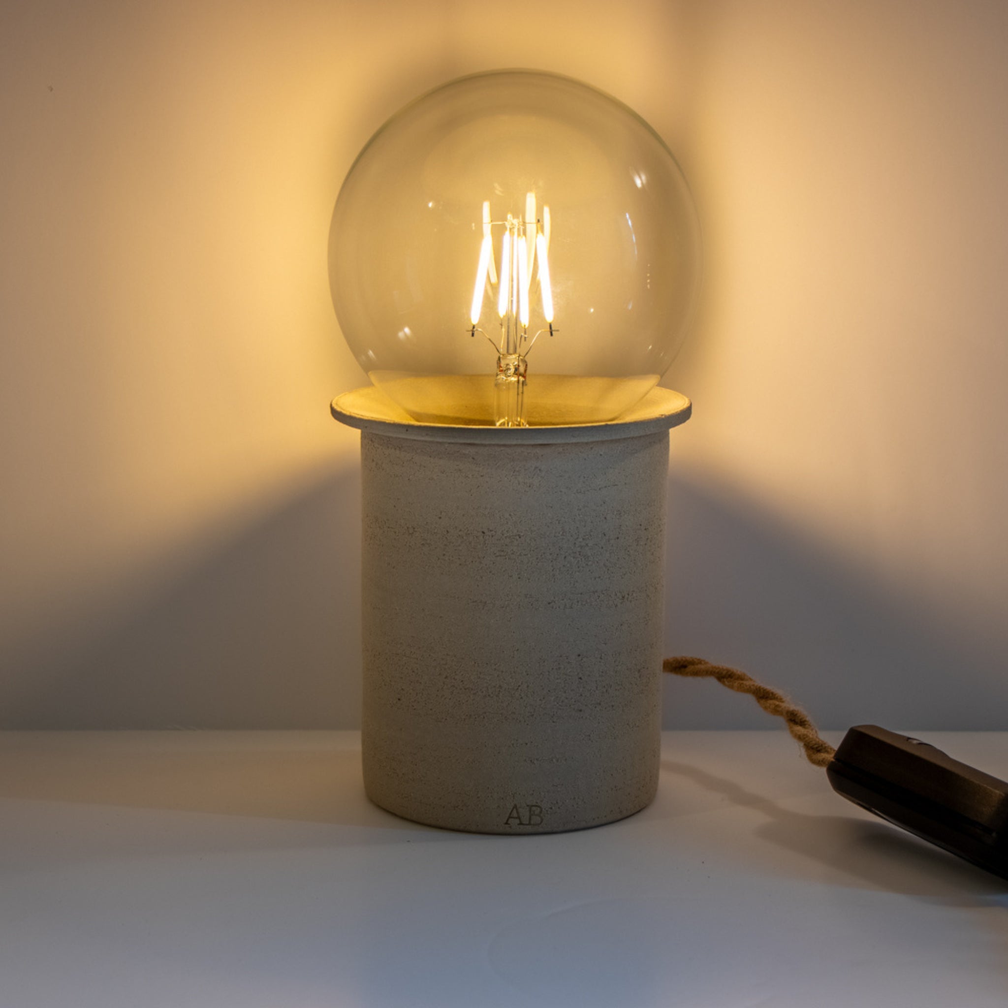 De AB Ceramics LED Lamp Dimmer heeft een grote glazen lamp in vintage stijl op een cilindrische beige keramische voet. Een handgemaakt touwachtig snoer en een donkerhouten schakelaar zijn zichtbaar en stralen warm licht uit tegen een effen achtergrond.