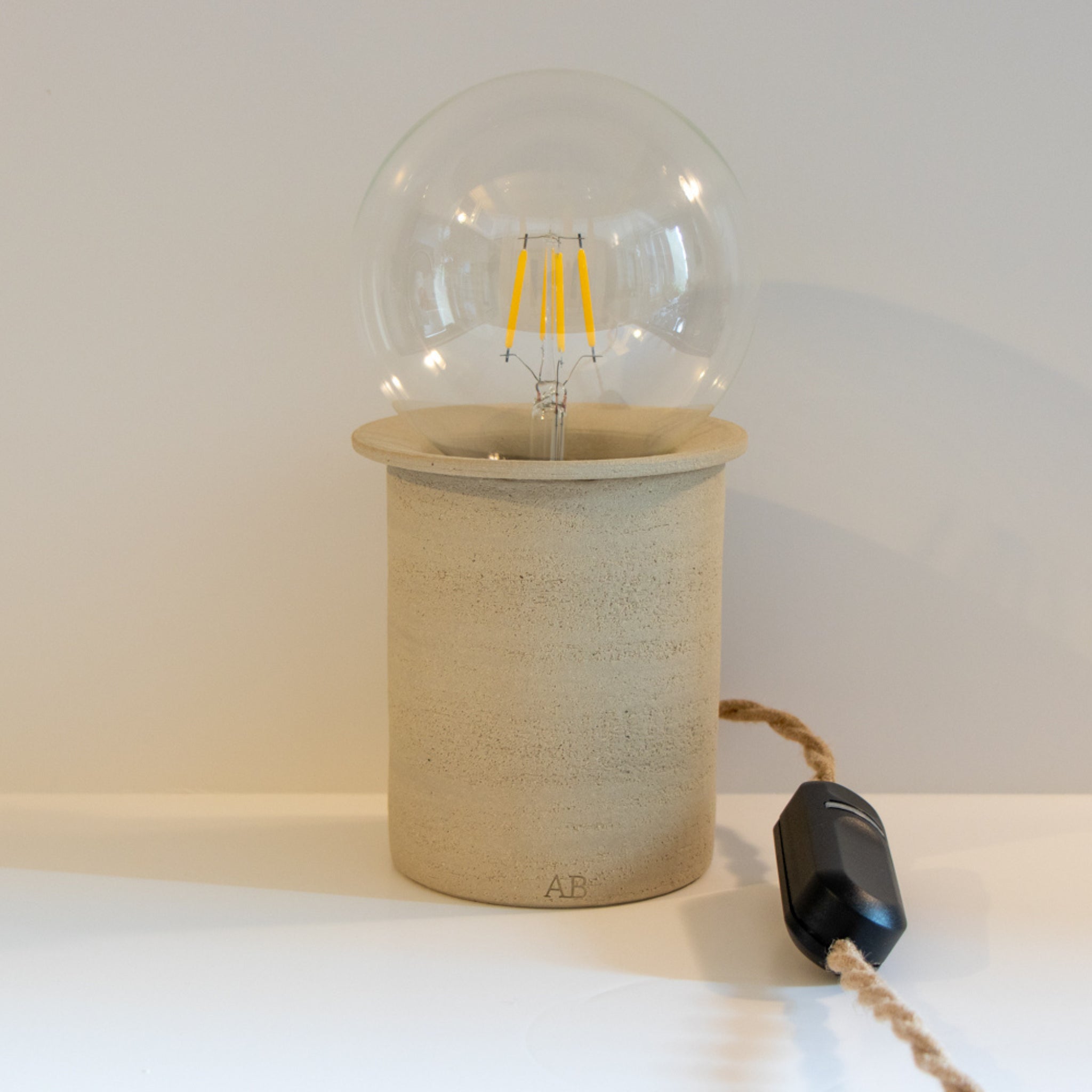 De AB Ceramics LED Lamp Dimmer is een minimalistische, handgemaakte lamp met een ronde heldere lamp en cilindrische beige keramische voet. De lamp heeft een bruin stoffen snoer en een zwarte inline schakelaar, gemaakt door AB Ceramics.